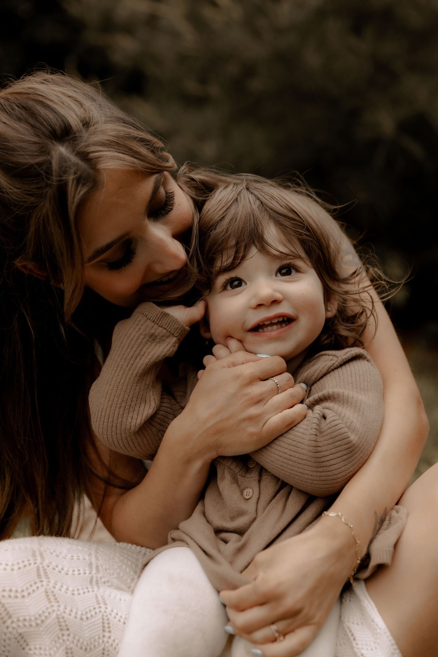 Mother and young child share loving embrace while wearing matching beige sweaters in moody outdoor portrait.