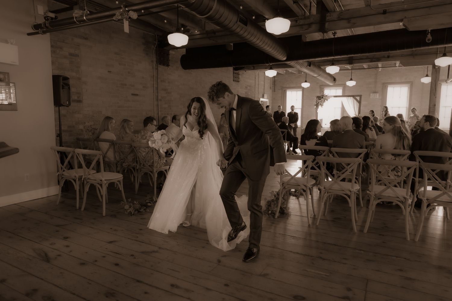 A candid wedding moment photographed in sepia showing movement and emotion in an industrial-style venue.