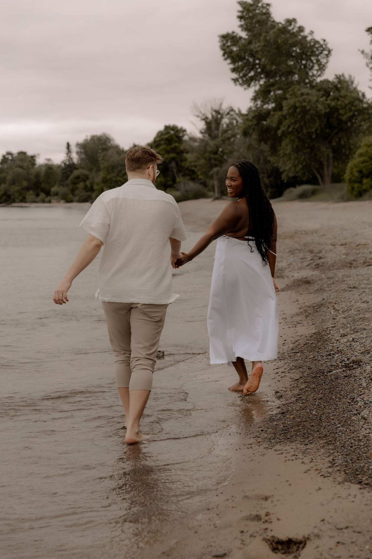 A couple holds hands while walking barefoot along a misty beach shoreline at dusk.