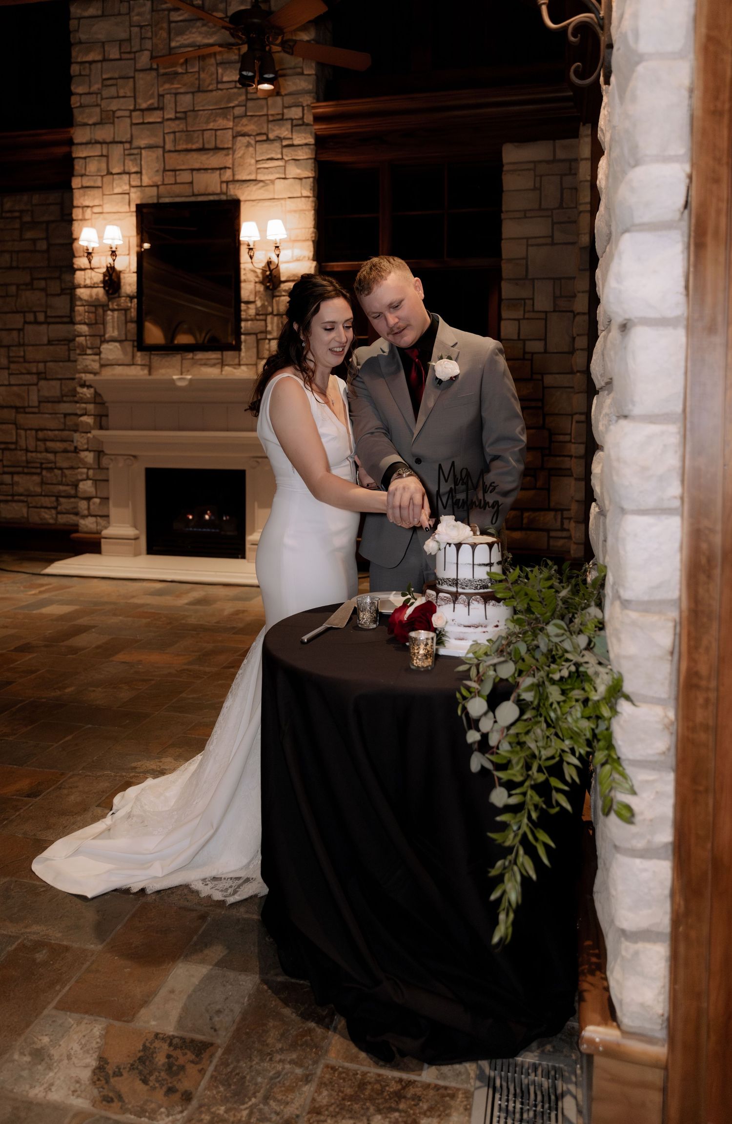 Bride and groom cutting their wedding cake beside a stone fireplace.