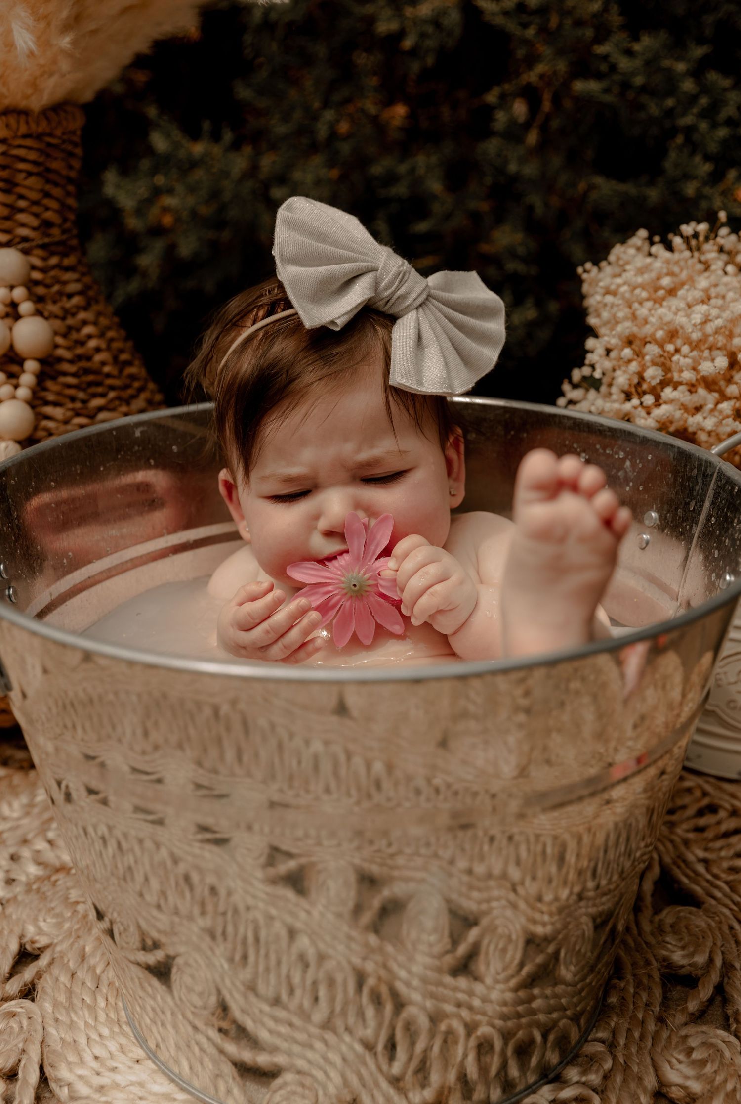 A small baby wearing a beige bow sits in a vintage metal washtub while playing with a pink flower in a garden setting.