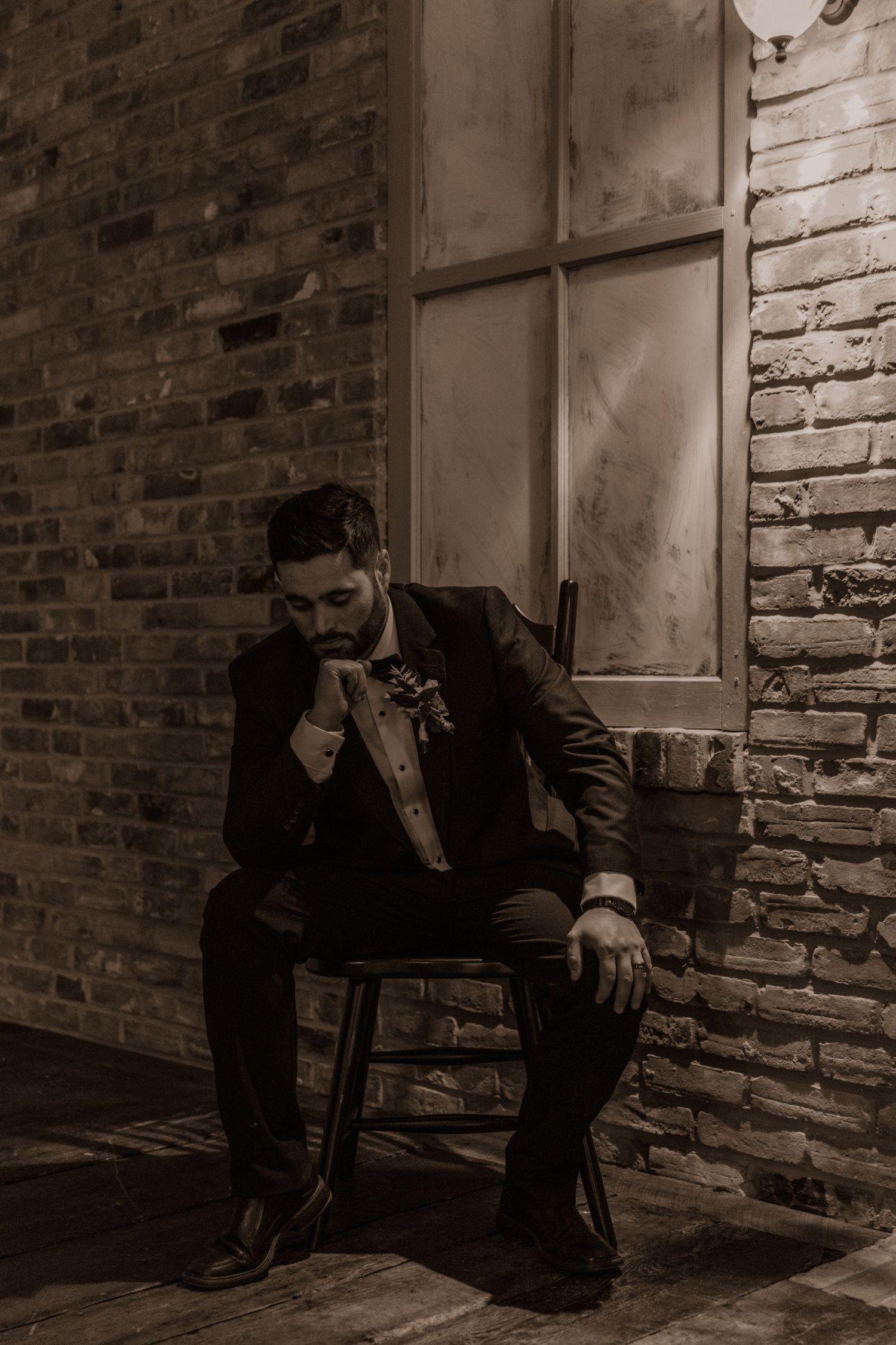 Pensive groom seated on wooden chair by vintage window and exposed brick wall.