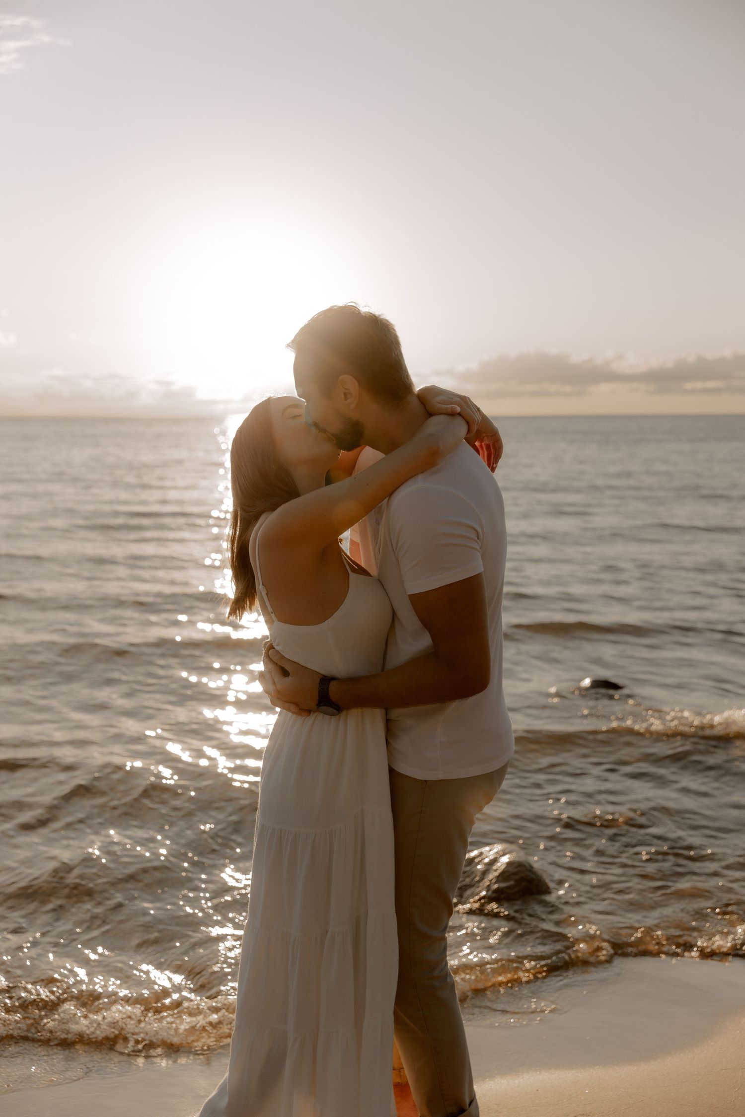 A tender beach embrace at sunset with warm golden light illuminating the waves and sand.