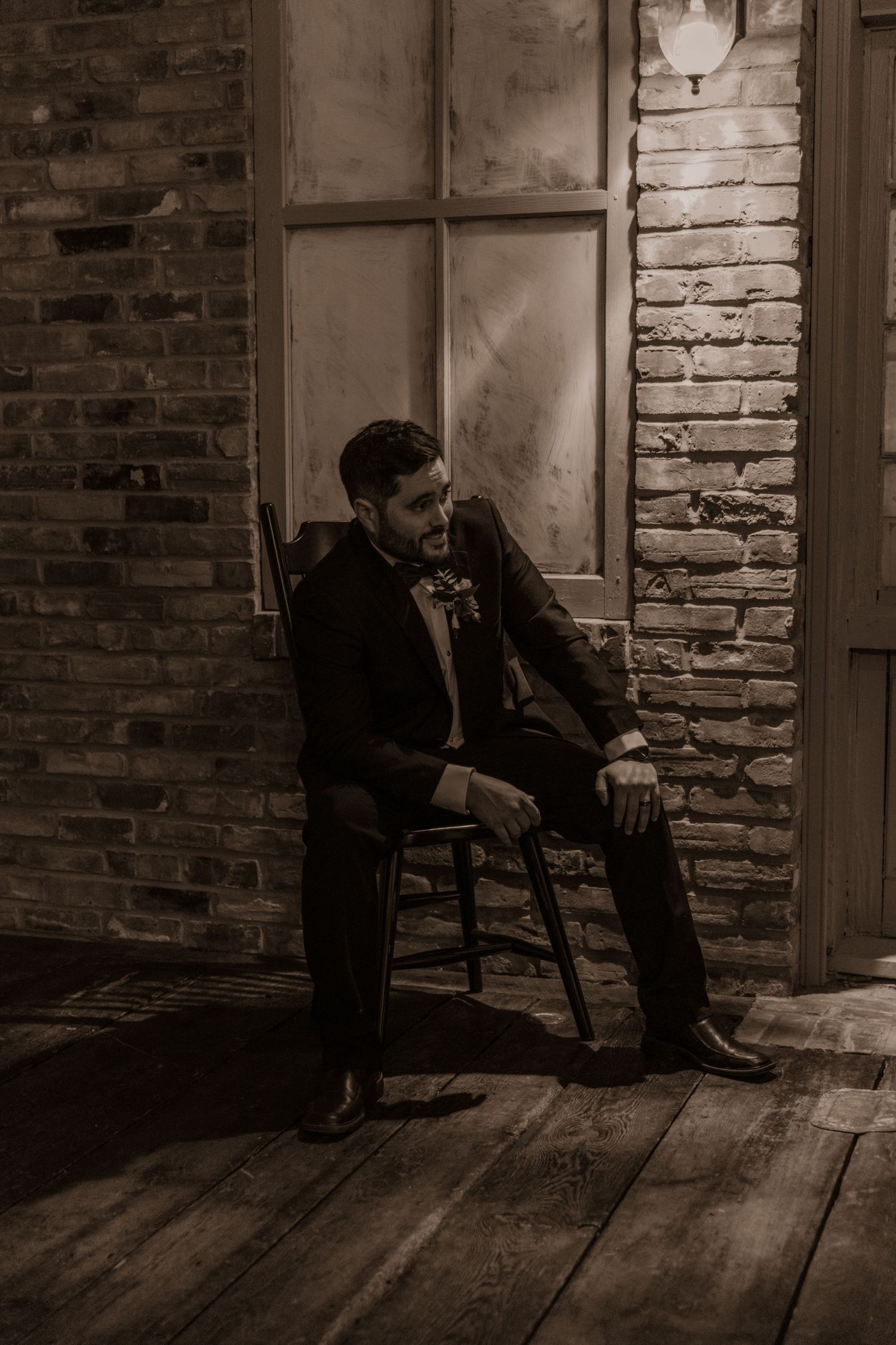 Groom in formal suit sitting alone on wooden chair against brick wall with window.