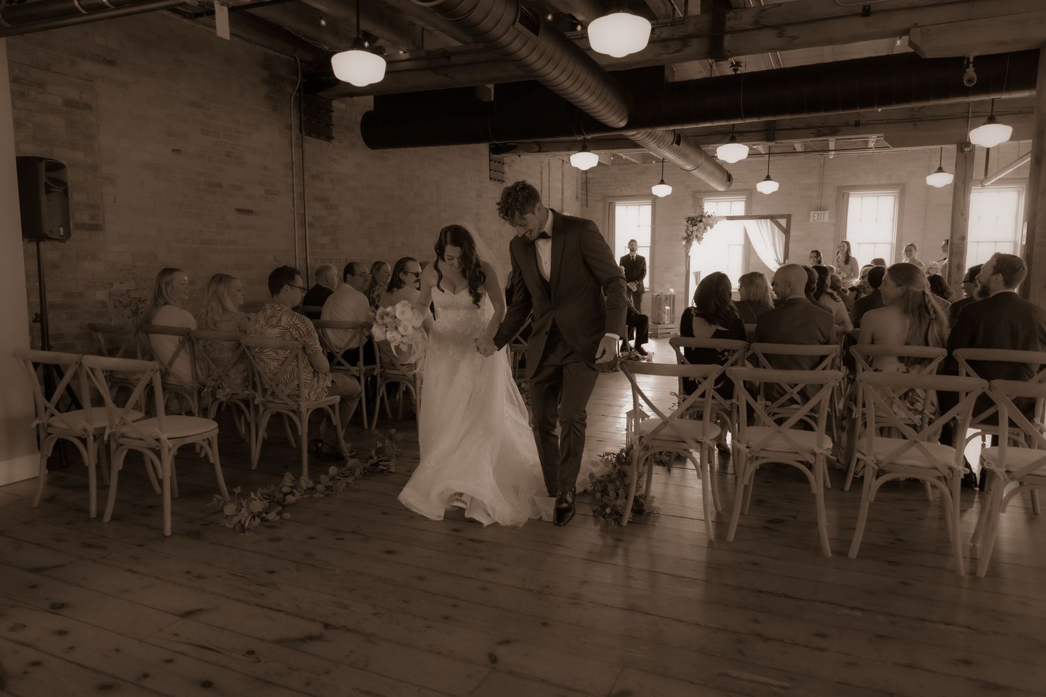 Wedding ceremony in rustic venue with wooden chairs and exposed brick walls.