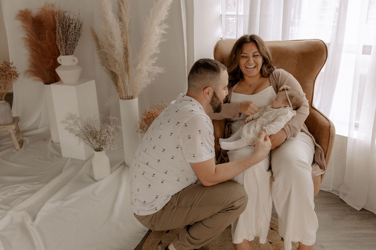 Family shares tender moment together while sitting in leather armchair near window with dried floral decor.