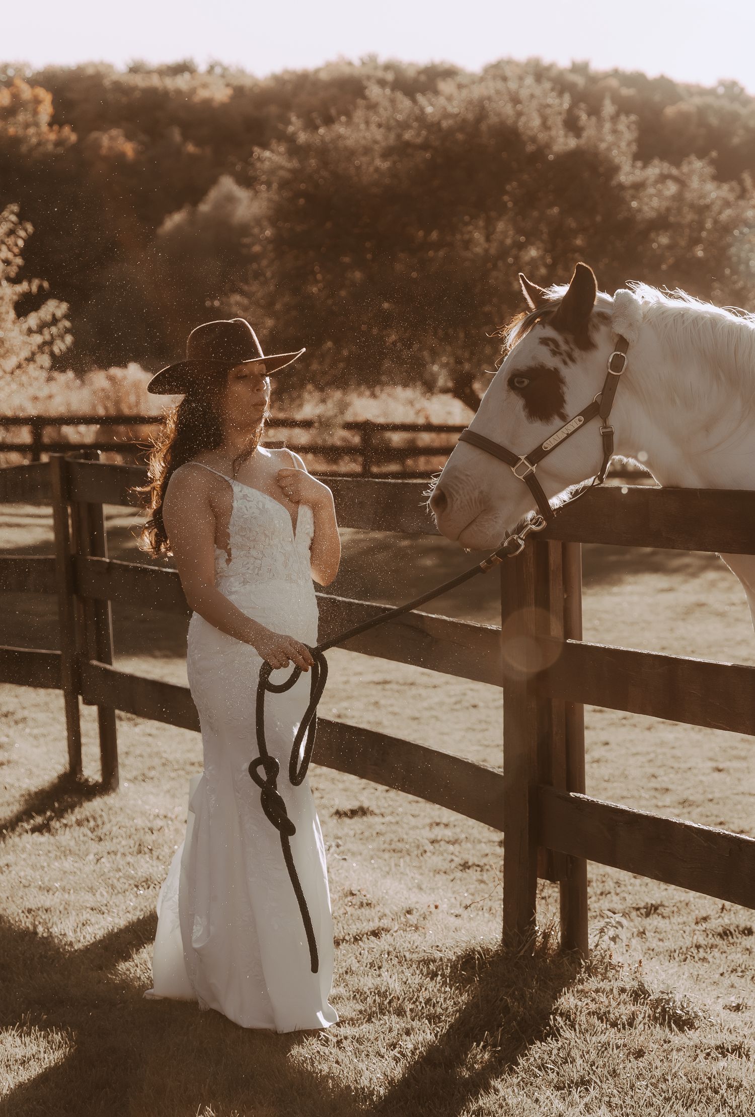 Someone in a wedding dress and cowboy hat interacts with a white horse at a rustic fence.