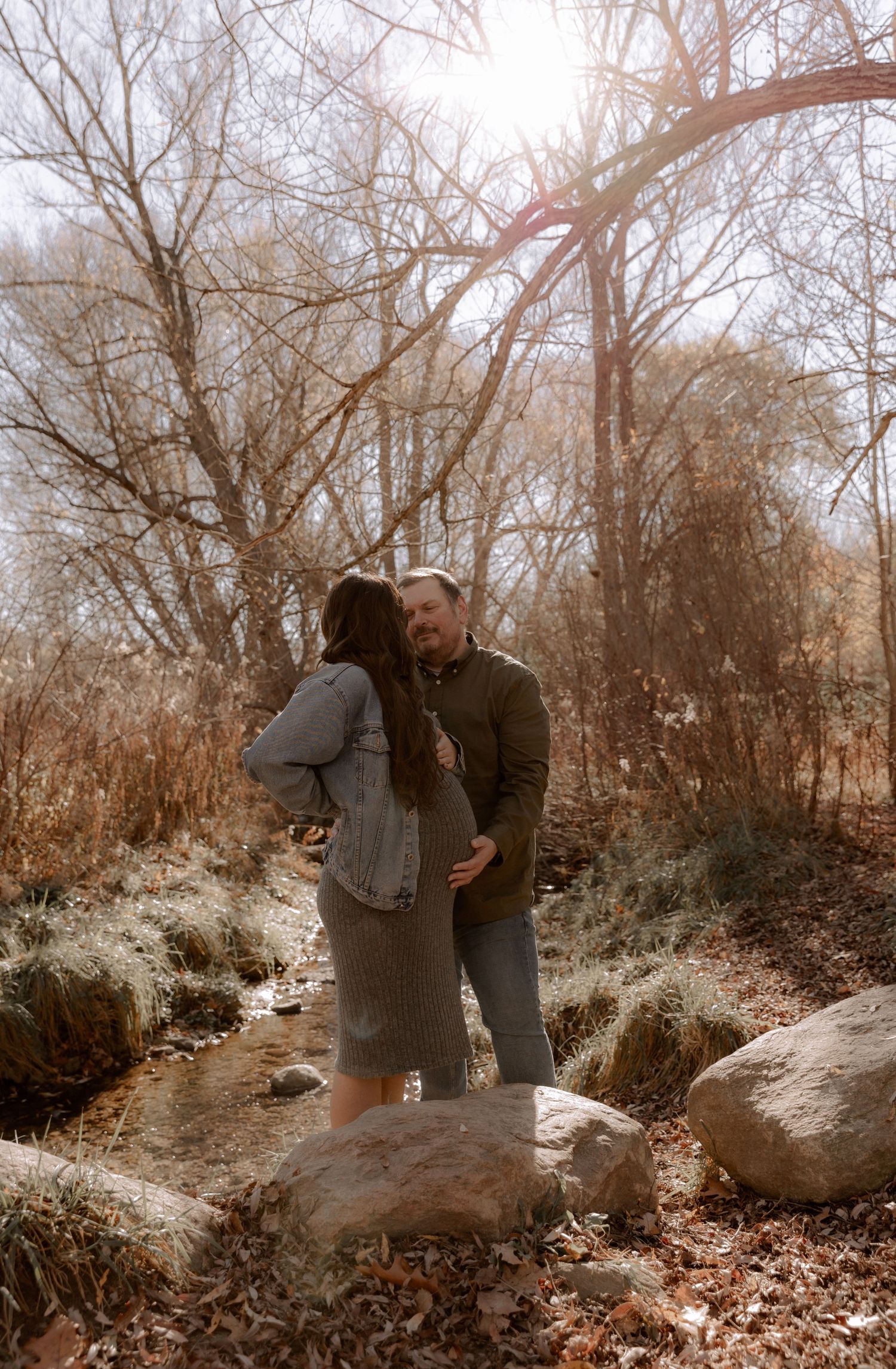 A couple embraces on rocks by a stream with sunlight filtering through bare winter trees above.
