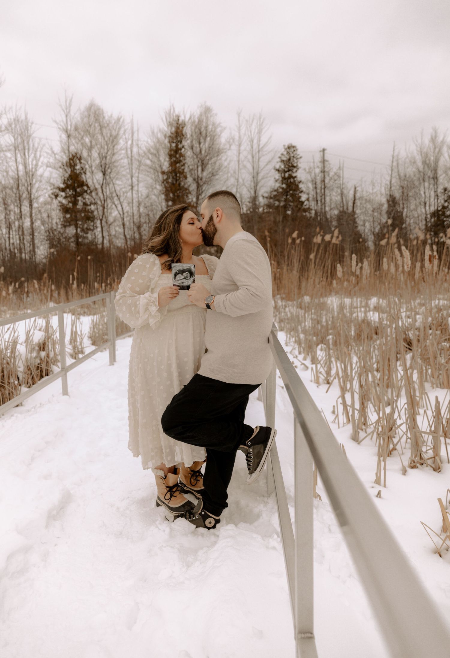 A couple shares a romantic moment on a snow-covered bridge surrounded by winter trees in a moody atmospheric setting.