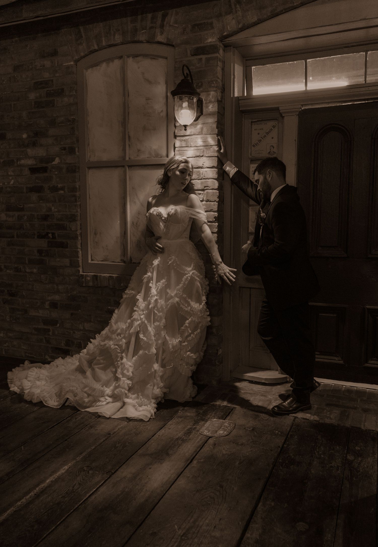 Vintage sepia photo of a couple in wedding attire interacting near an old wooden door and brick wall.