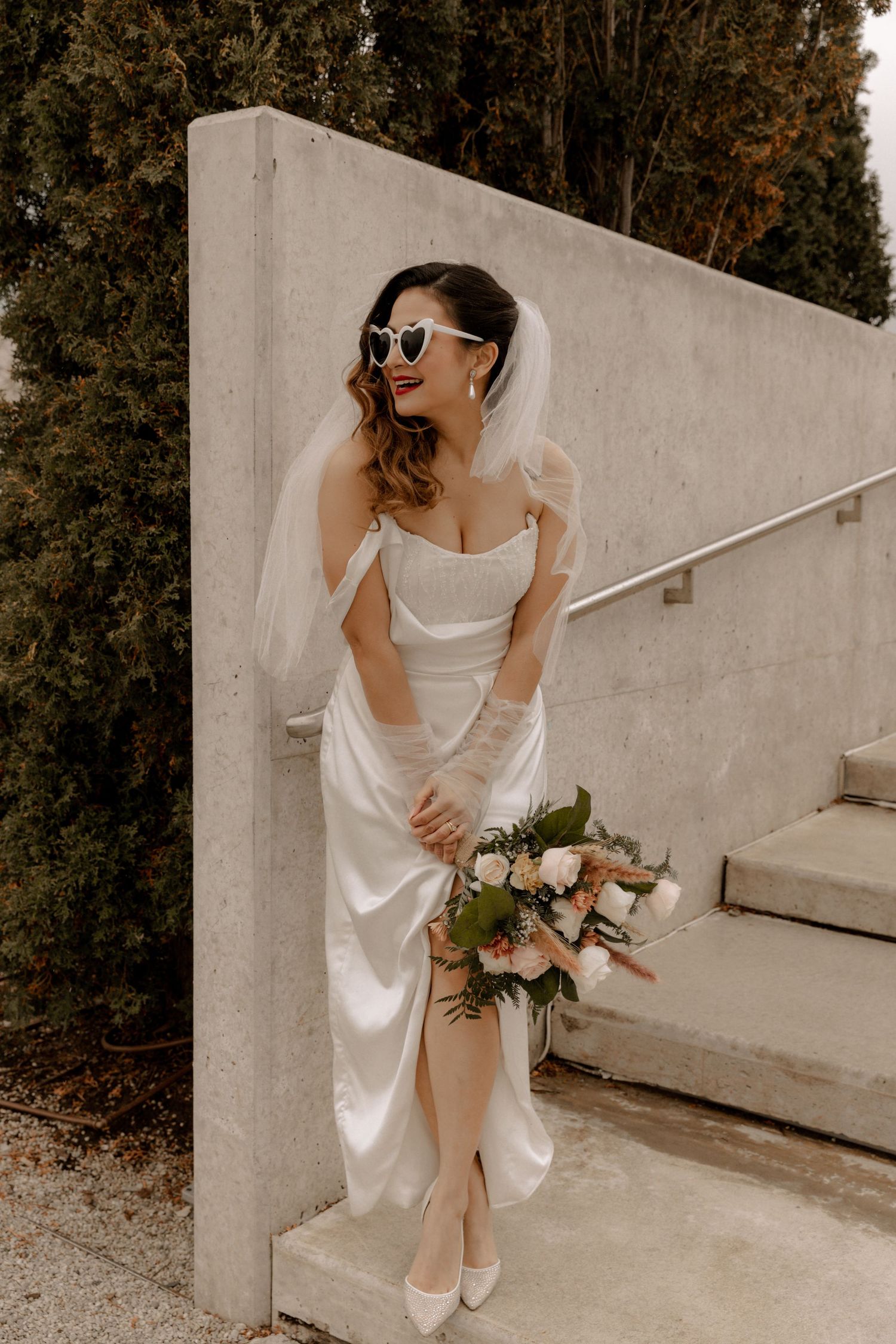 Elegant bride in silk dress holds bouquet against concrete wall.