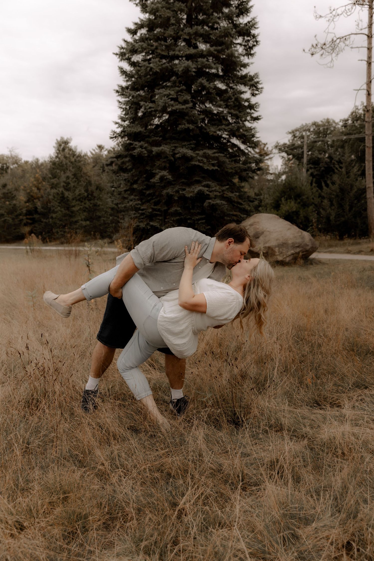 A romantic dance dip pose captured in a rustic field setting with evergreen trees.