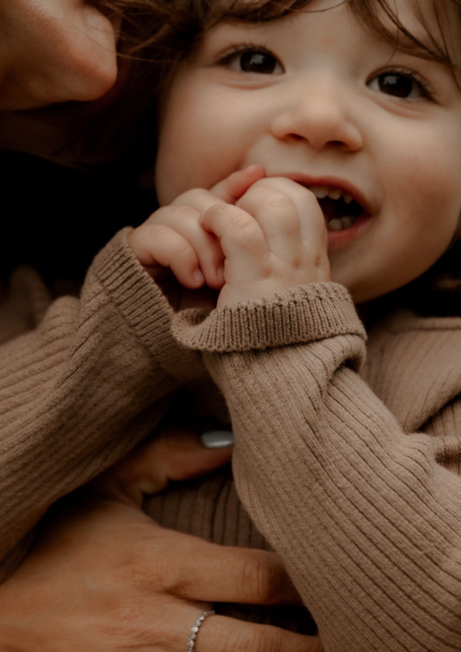 Close-up of a small child wearing a cozy brown knit sweater while smiling and touching their face.