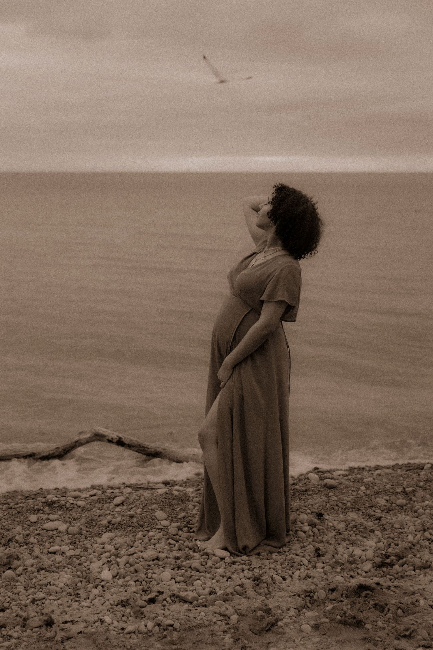 Figure in a flowing dress stands on a seaside cliff looking up at the sky in a dreamy sepia-toned vintage photograph.