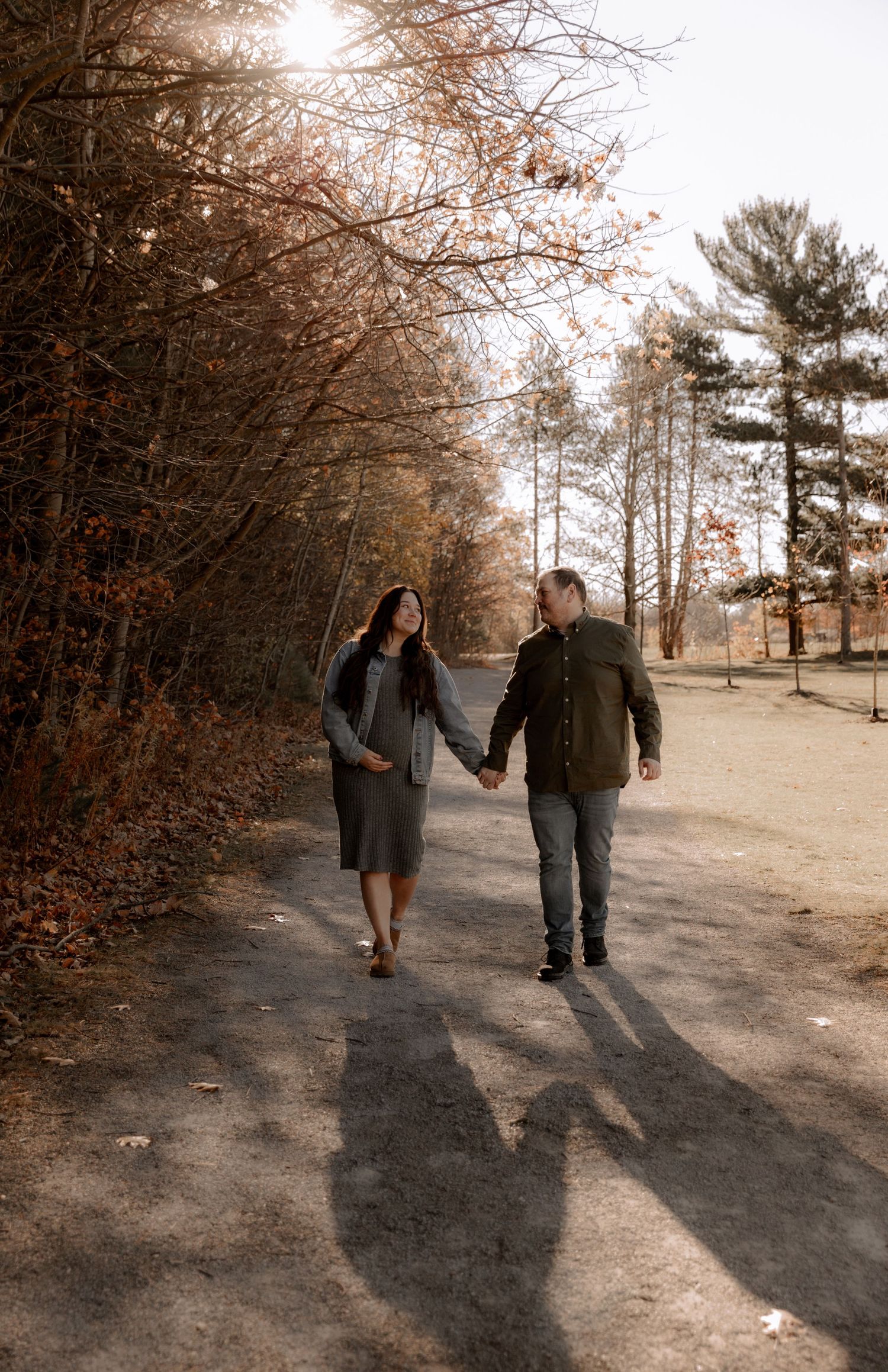 A couple holds hands while walking down a sunlit autumn path surrounded by bare trees and fallen leaves.