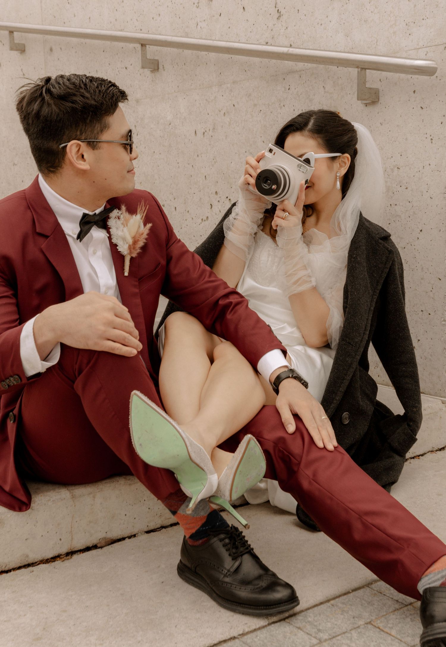 A stylish wedding couple sits playfully on steps with vintage camera and burgundy suit.