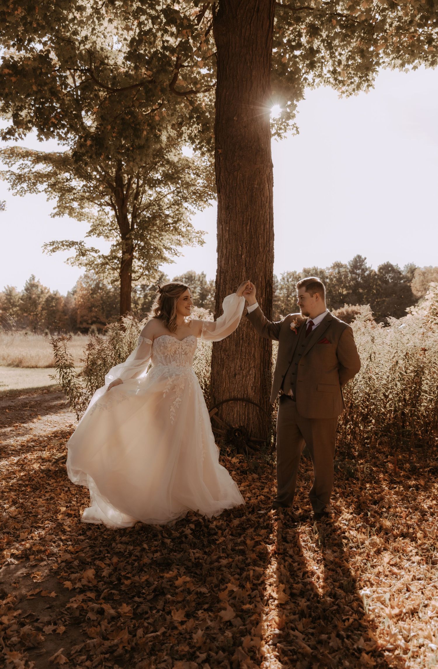A couple in wedding attire playfully dance around a tree trunk in a dreamy sepia-toned forest setting.