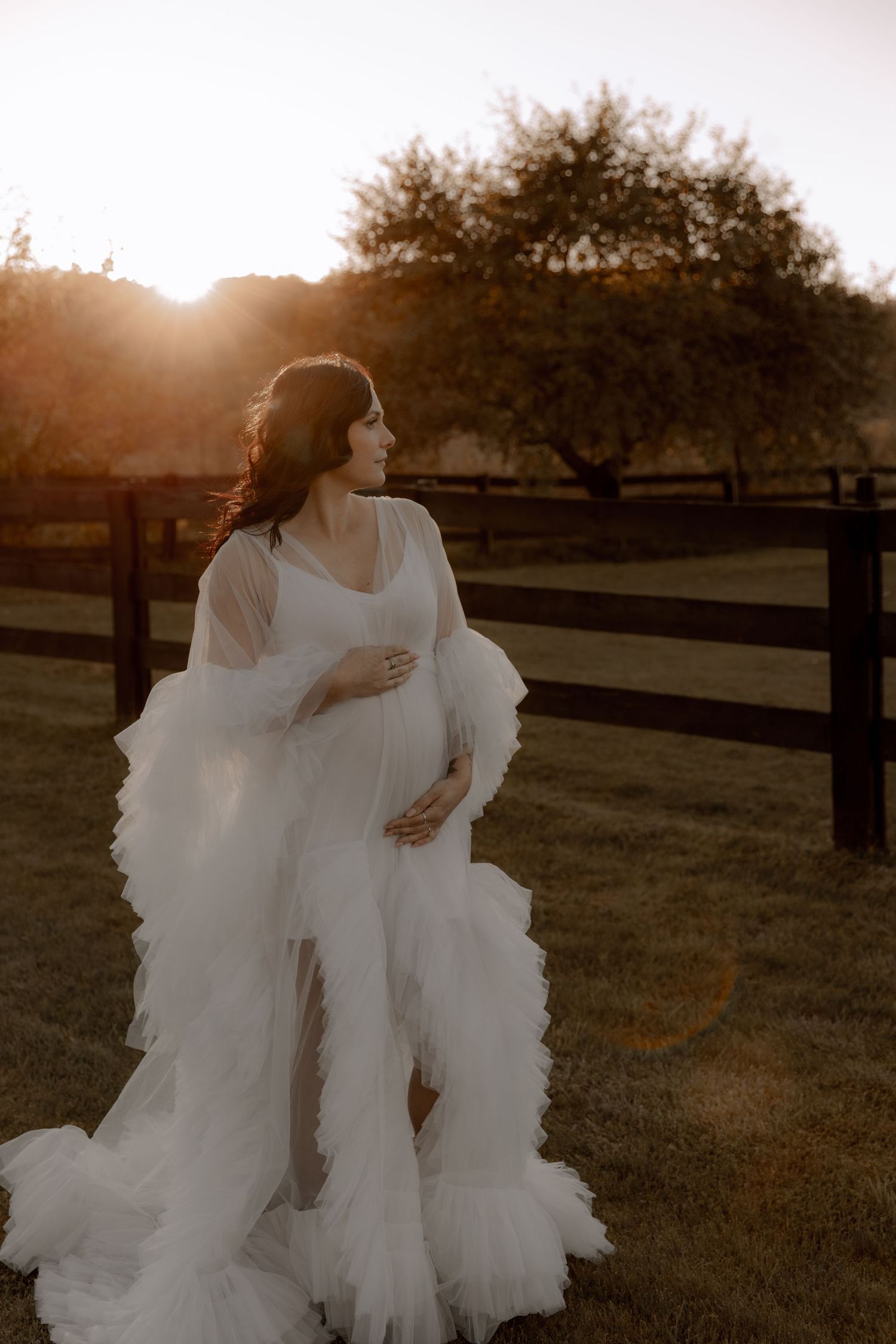 A maternity photoshoot at sunset features an ethereal white ruffled gown against a rustic wooden fence backdrop.