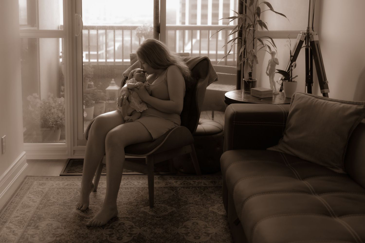 Figure sitting in armchair by window in sepia-toned living room interior with couch and houseplant.