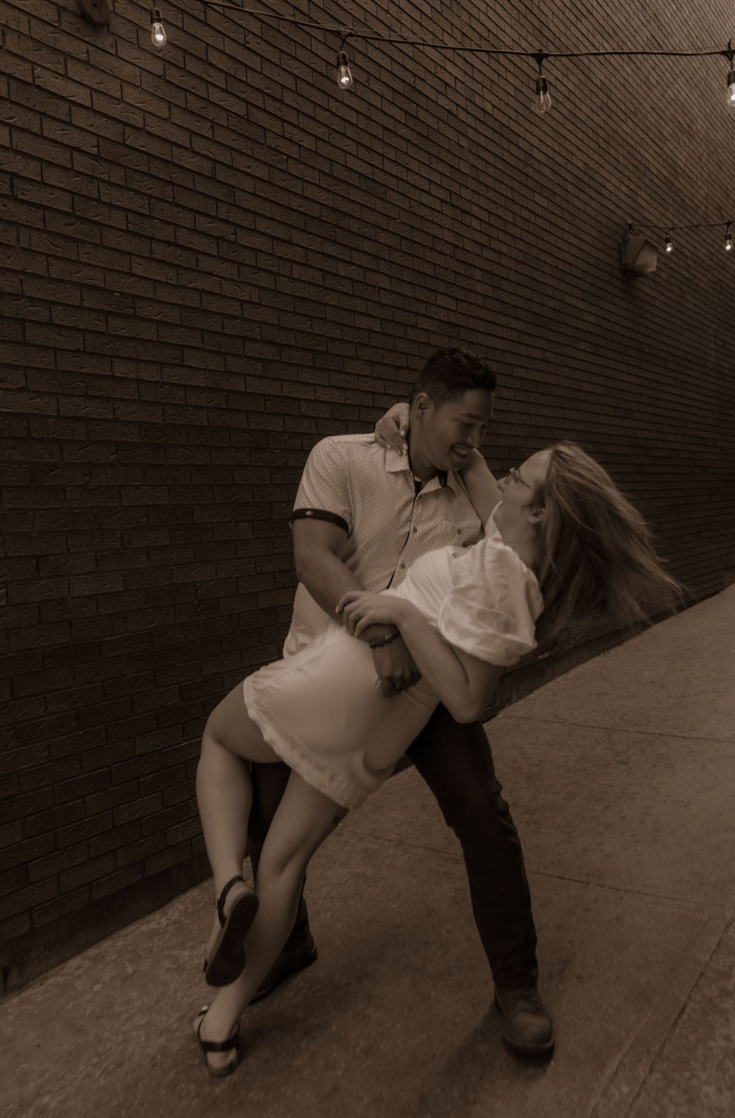 A romantic vintage-style dance dip pose against a brick wall with string lights overhead in sepia tones.