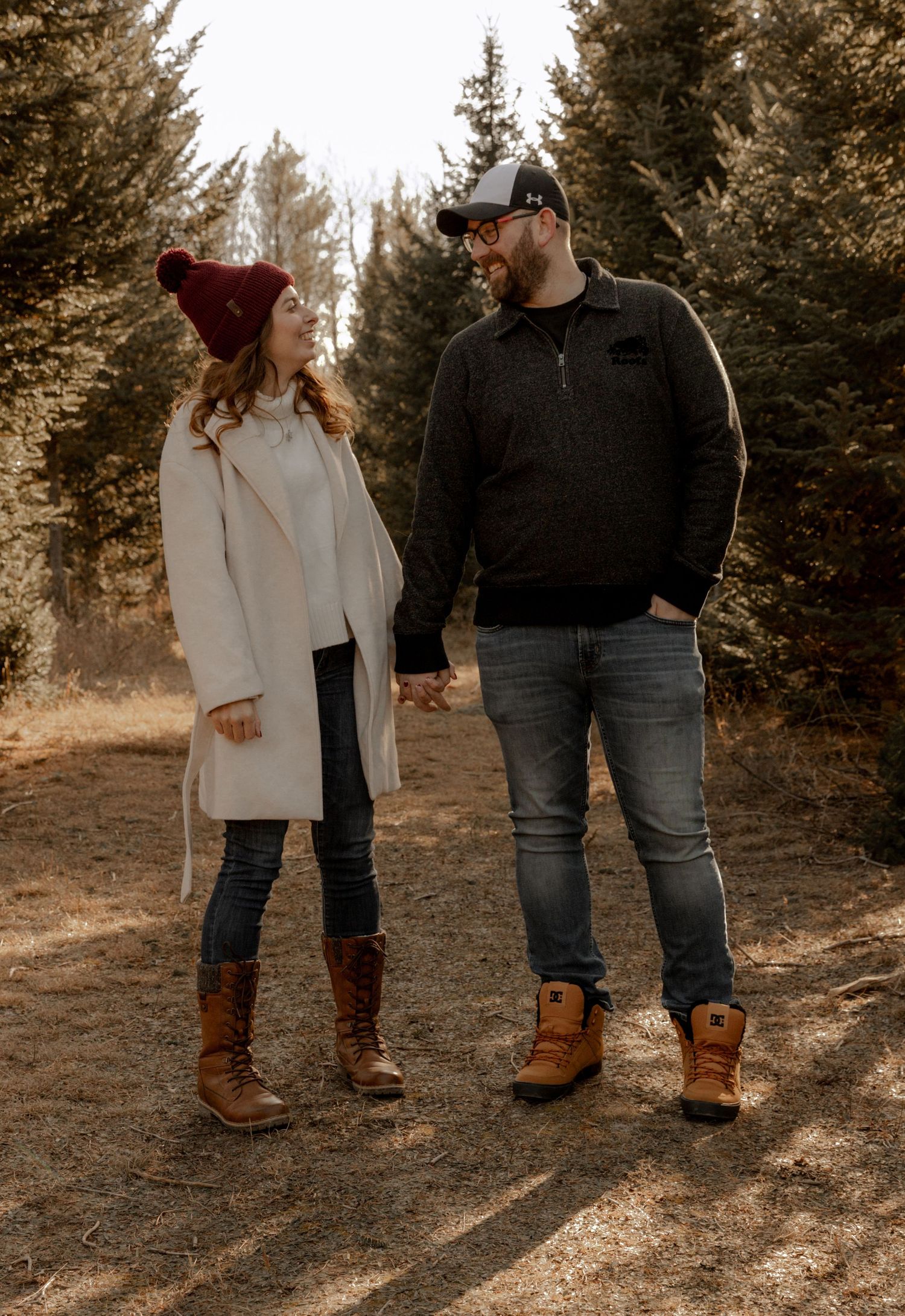 A couple holding hands while standing on a dirt path among evergreen trees wearing winter boots and coats.