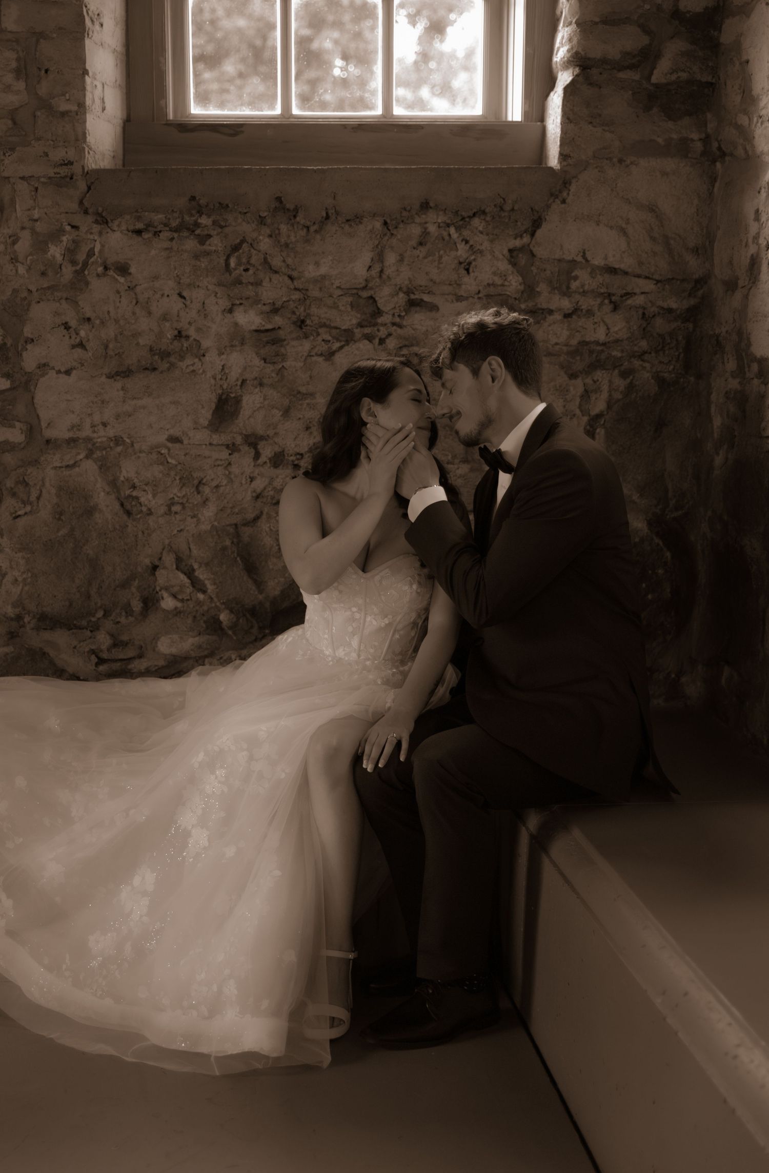 A sepia-toned wedding photo of a couple in an intimate moment against a stone wall with window light.