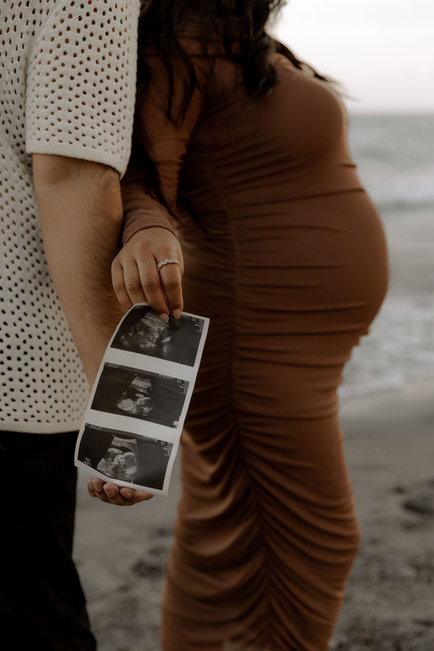 Close-up of hands holding ultrasound photos against a pregnant belly on a beach.
