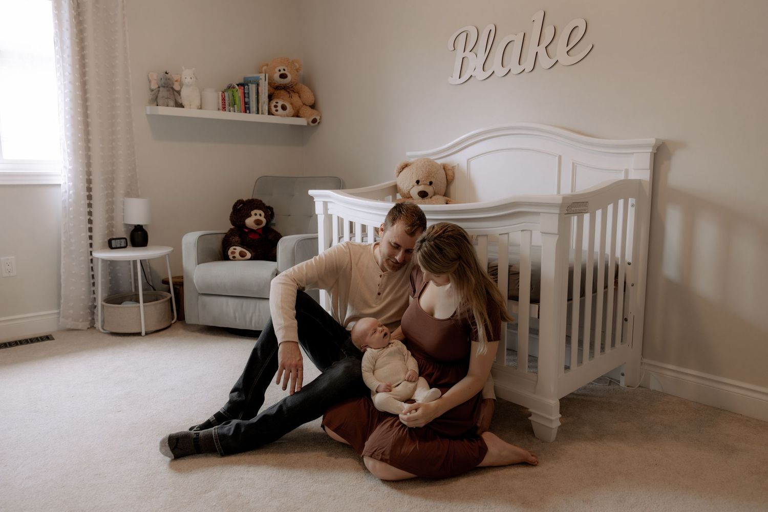 Cozy nursery room with white crib, stuffed animals on shelf, and grey armchair against neutral beige walls.