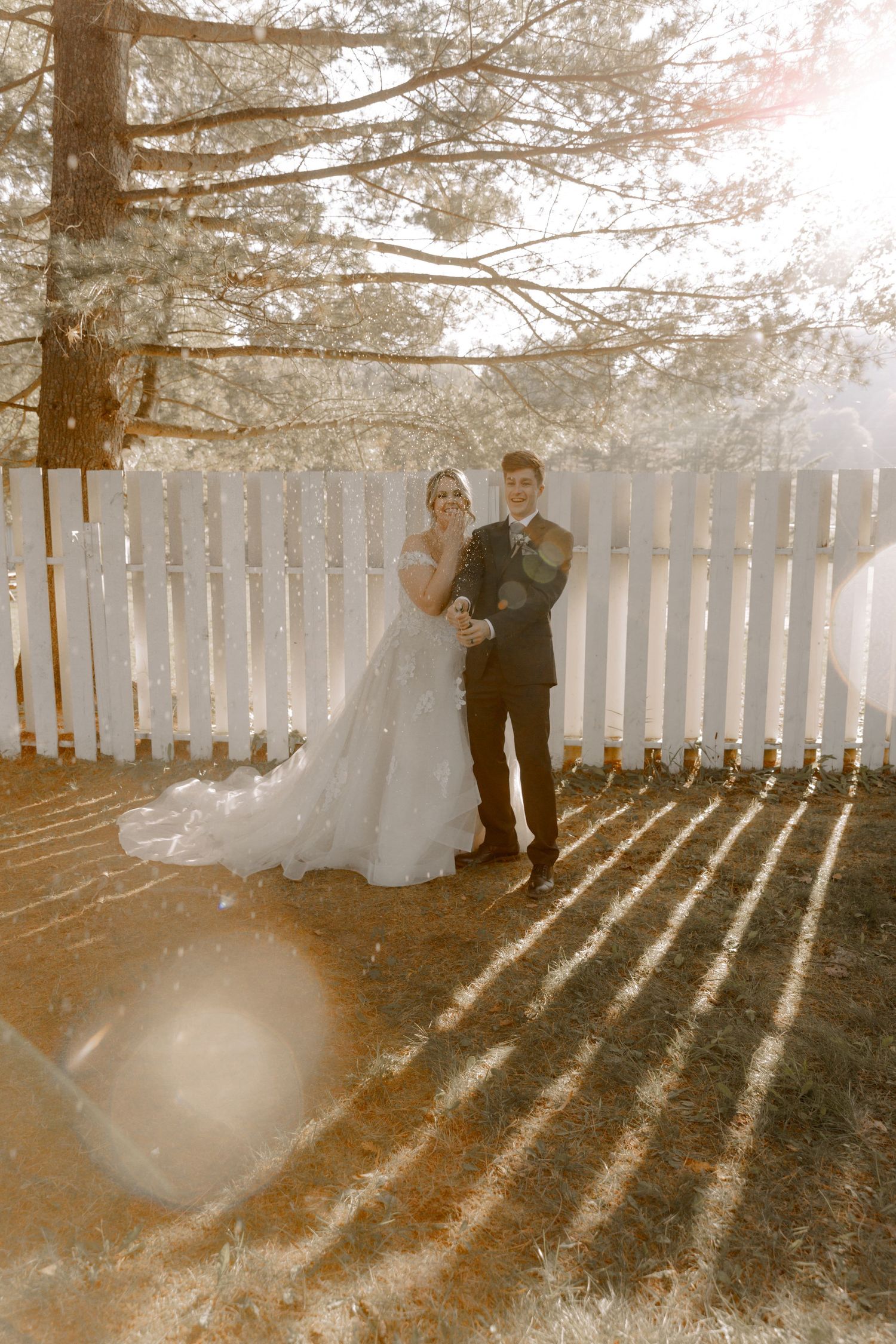 A sunlit wedding portrait against a white picket fence with dramatic shadows.