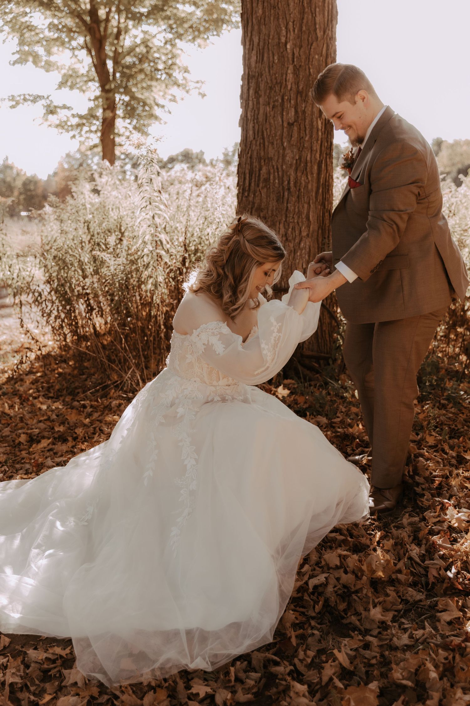 Romantic wedding moment captured as couple interacts beneath a towering tree with fallen leaves.