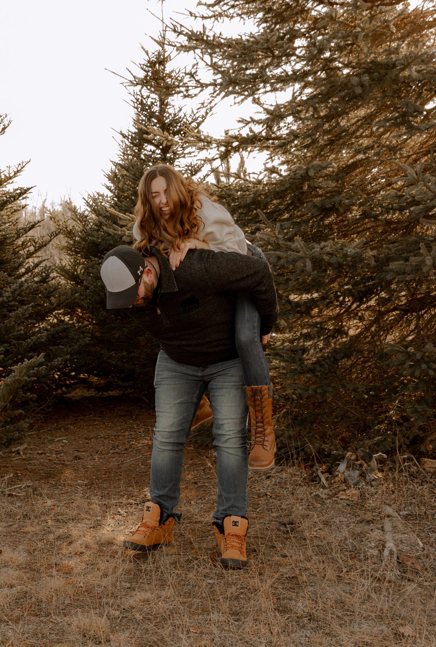 Two people having fun outdoors with a playful piggyback ride among evergreen trees at sunset.