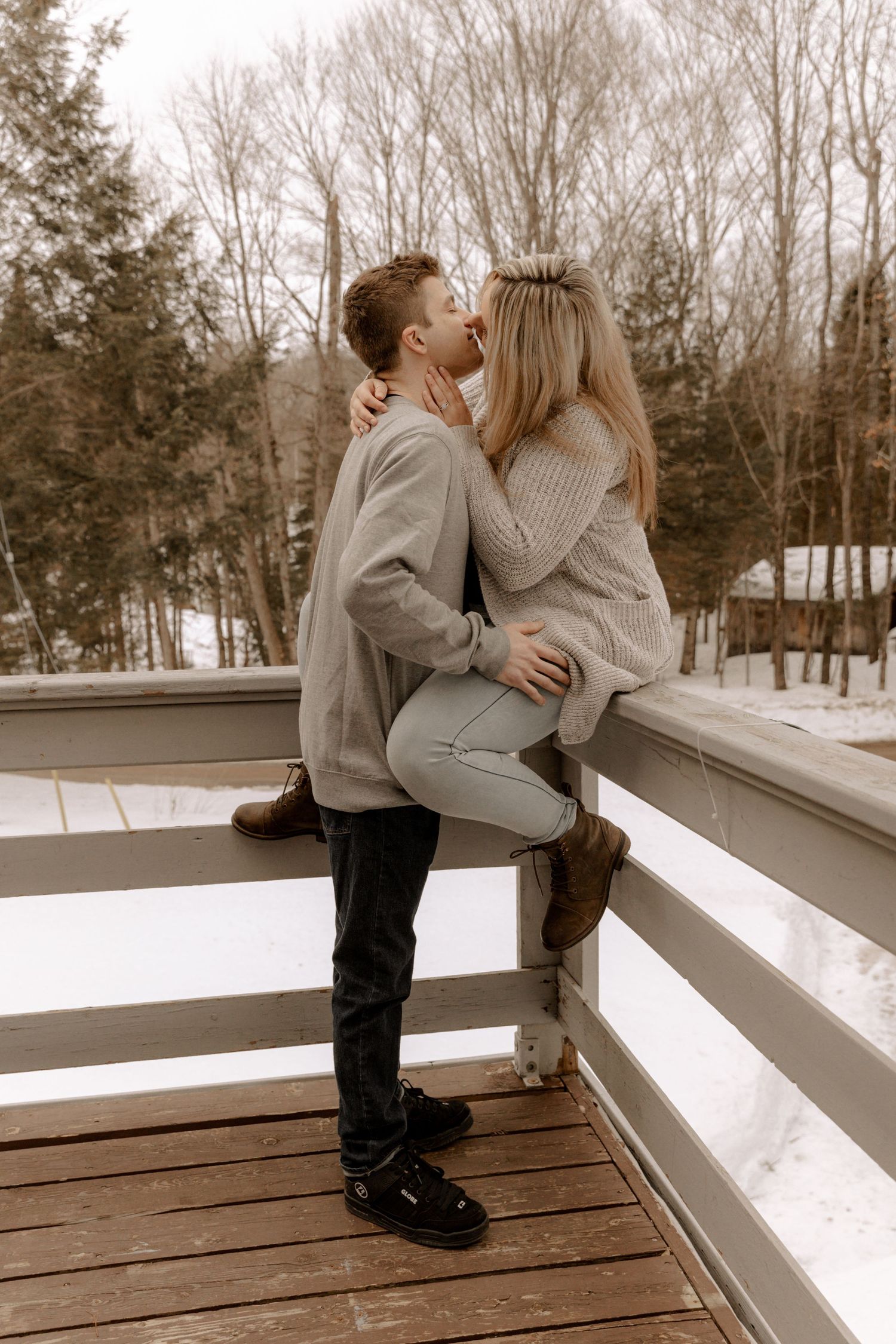 A romantic couple embraces on a wooden deck surrounded by snowy winter trees in a sepia-toned photo.