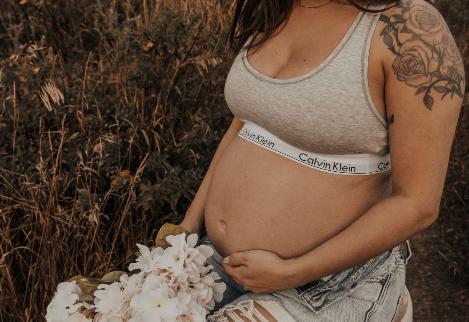 A pregnant person wearing a Calvin Klein sports bra sits amid dried foliage with white flowers resting nearby.