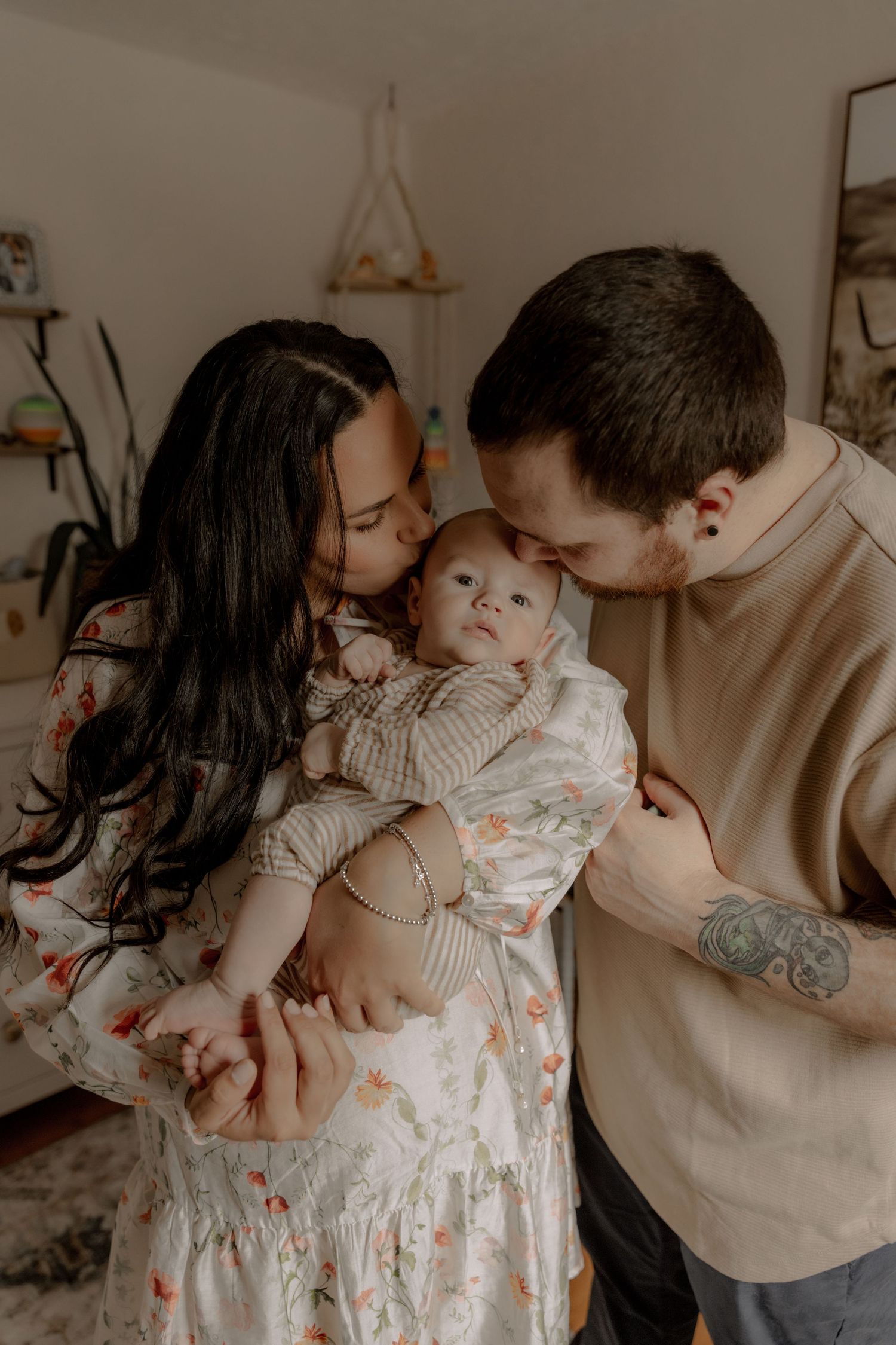 Intimate indoor family moment as parents tenderly embrace their baby in a softly lit room with minimal decor.