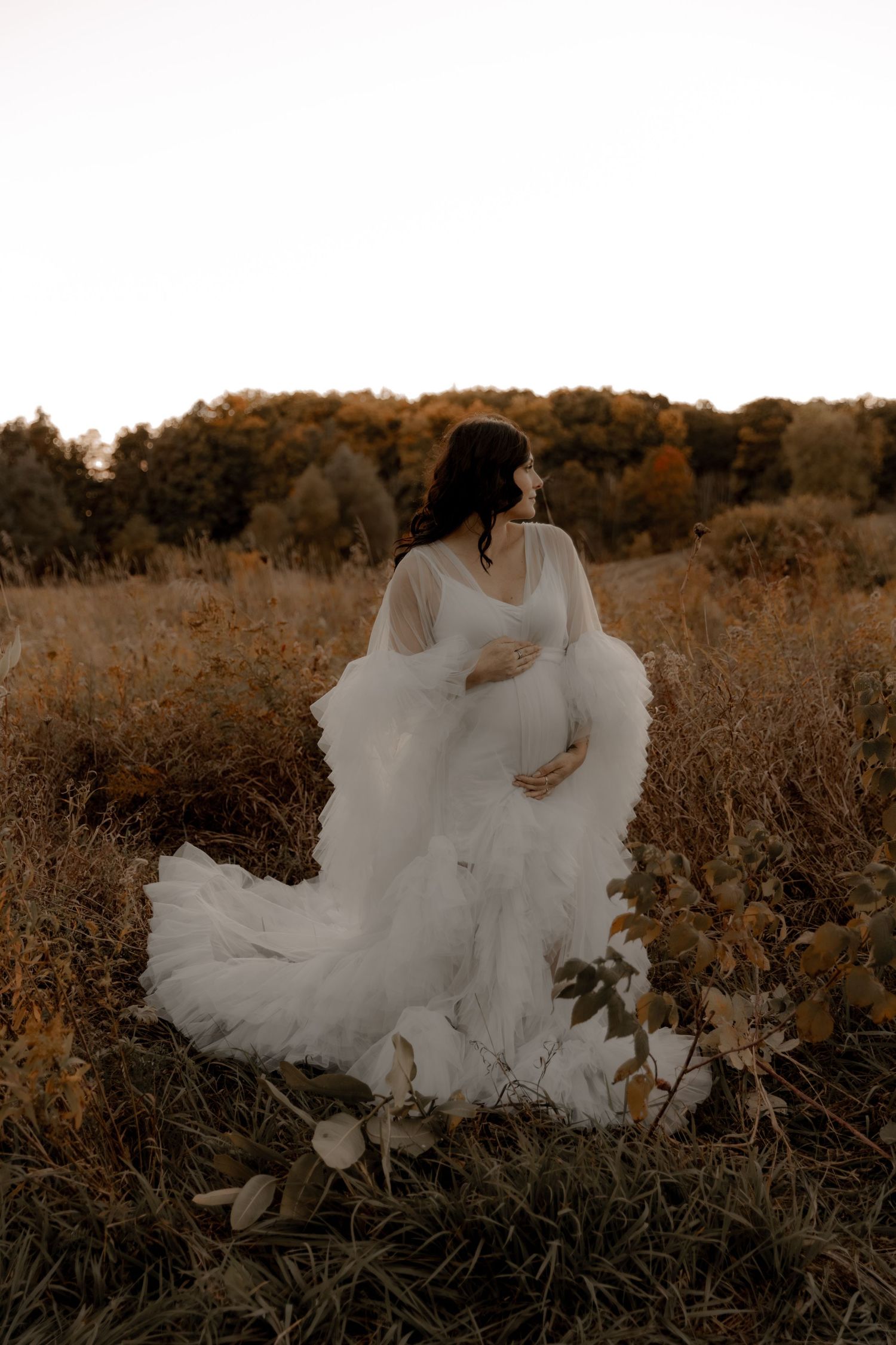 Ethereal maternity photo of woman in flowing white dress at golden hour.