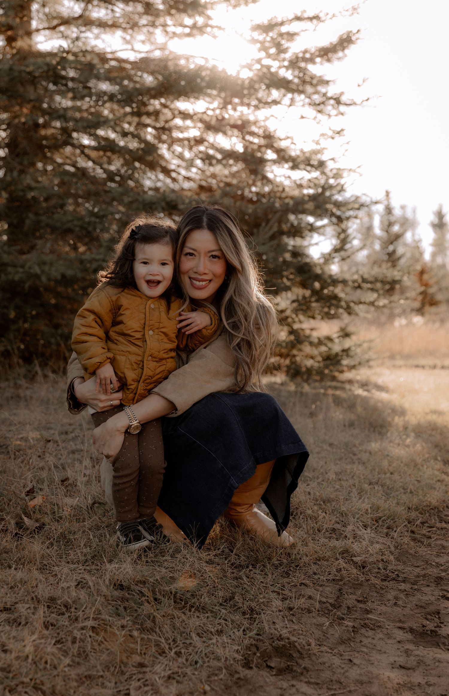 A parent and child wearing brown outfits pose together outdoors on a sunny autumn day with evergreen trees in background.