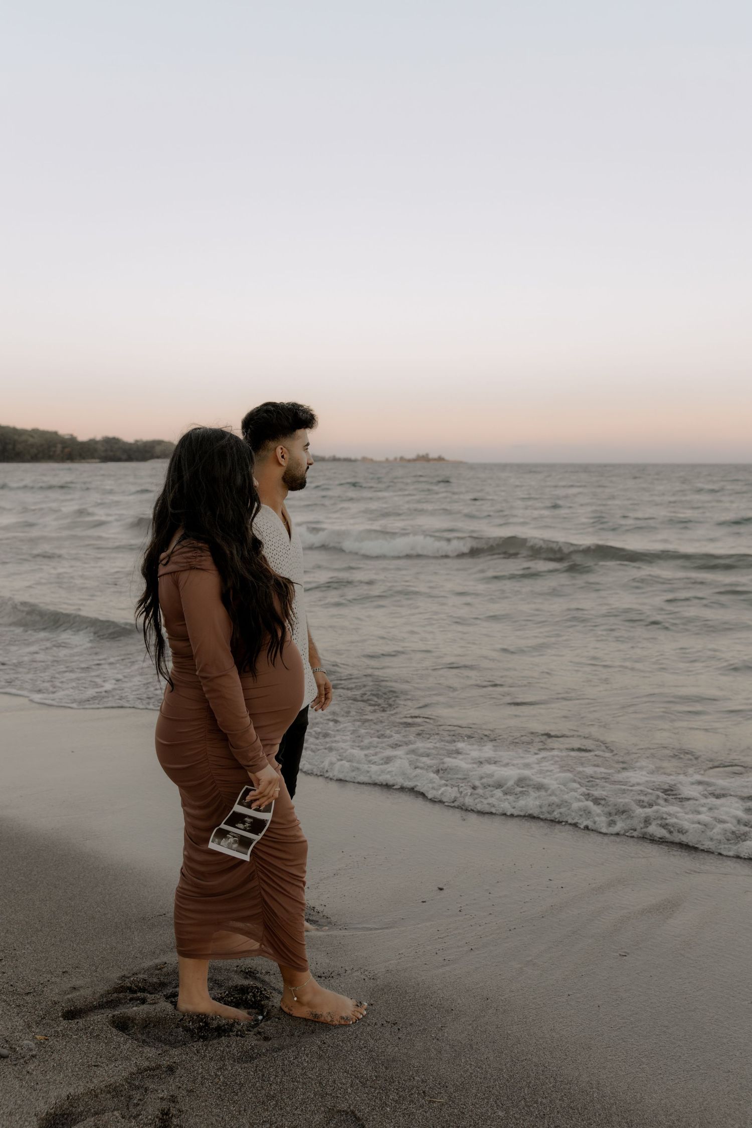A couple stands together on a beach at sunset watching the waves, while holding ultrasound photos.