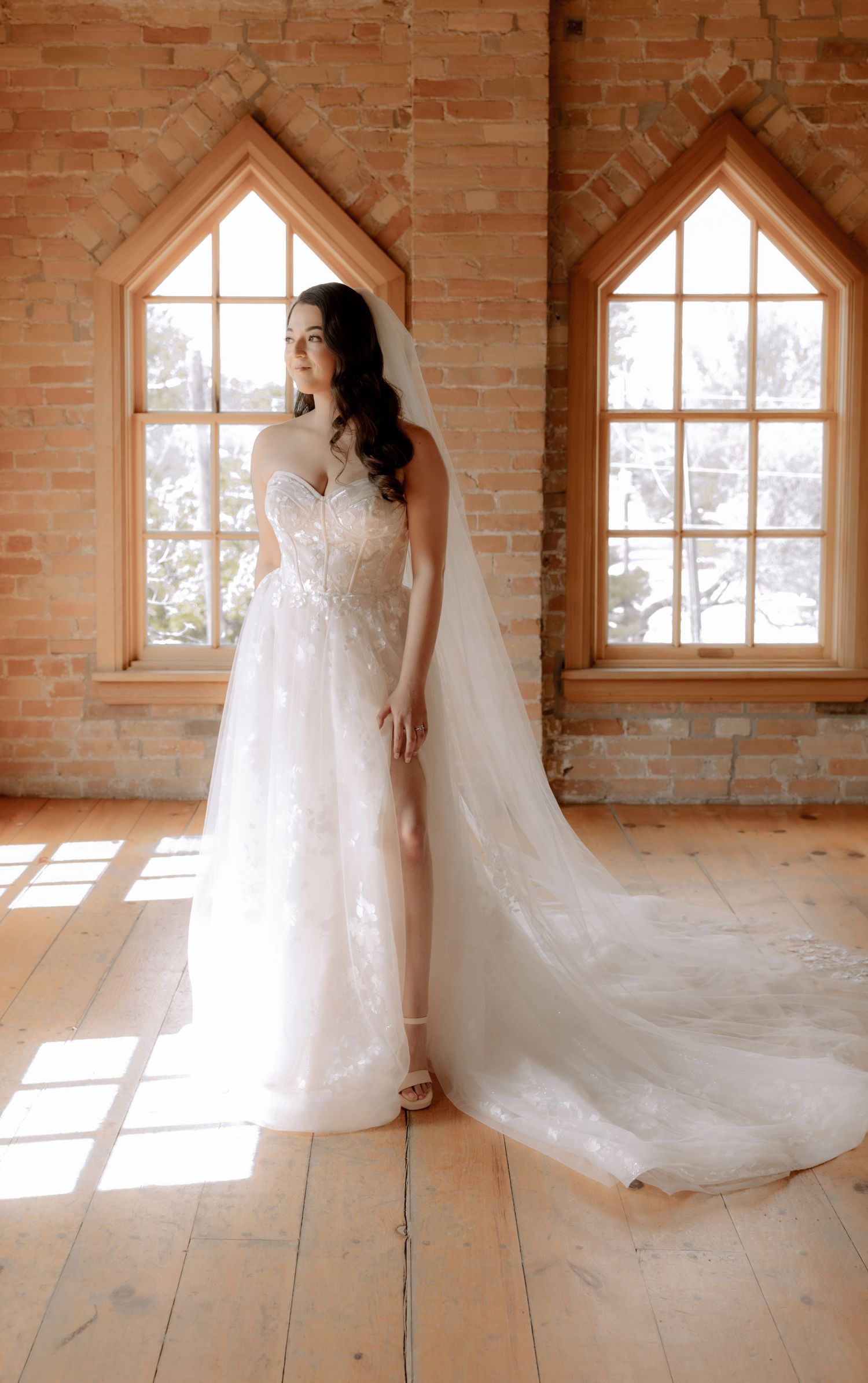 Stunning bridal portrait in a sunlit brick room featuring a lace gown with dramatic train.