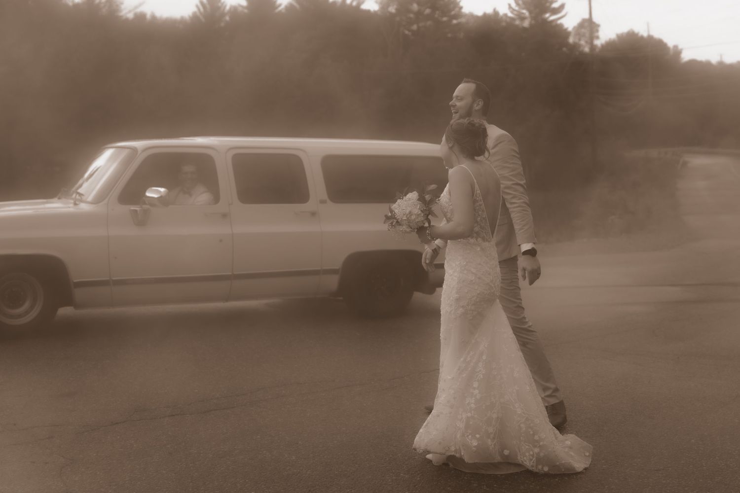Sepia photograph of a couple in wedding attire walking past a vintage suburban vehicle.