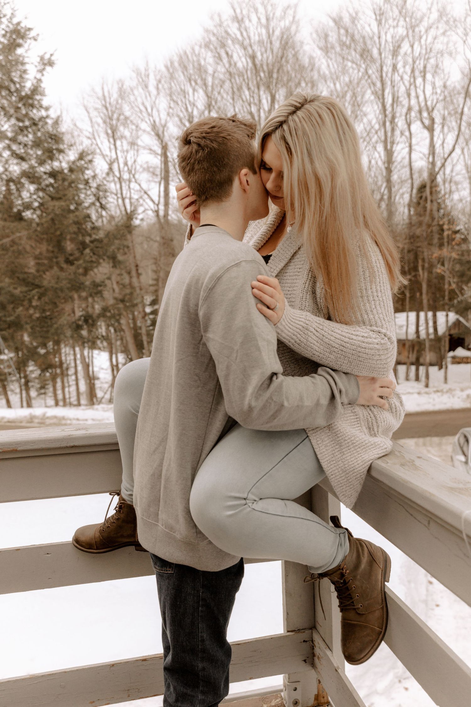 A romantic winter scene shows a couple embracing on a snowy wooden deck railing with bare trees in the background.