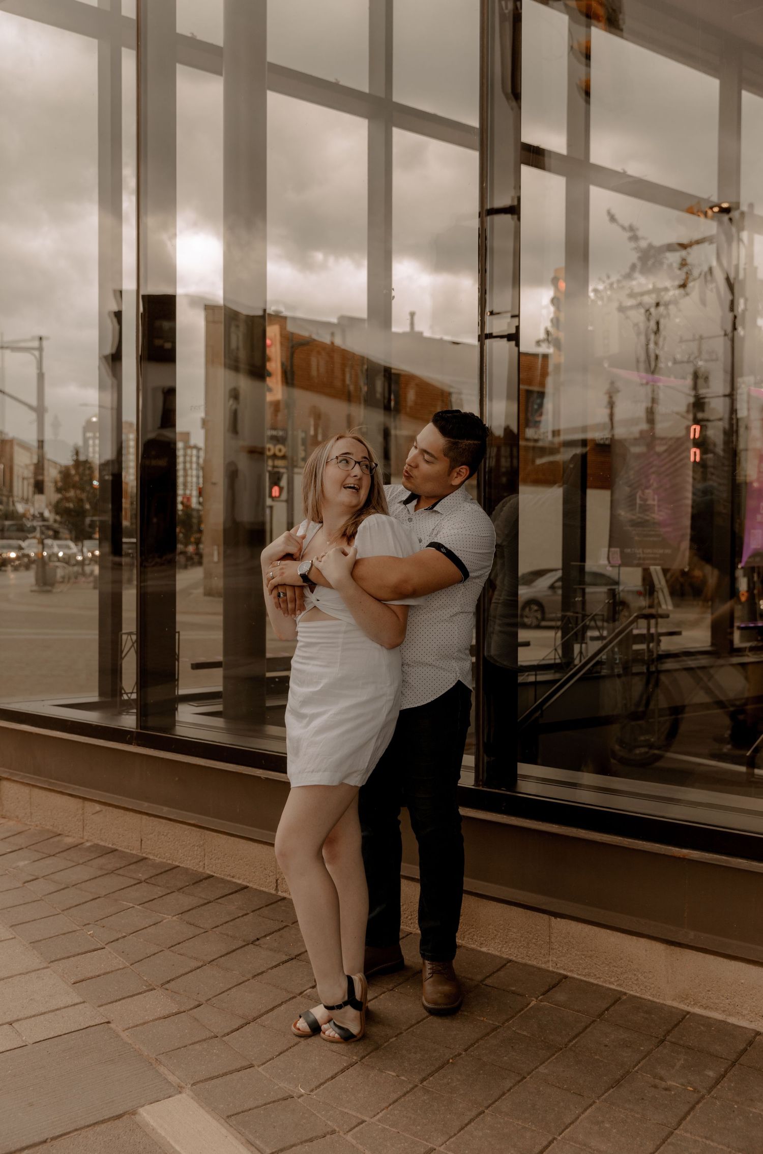 A couple shares an intimate moment outside storefront windows on an urban sidewalk during a cloudy day.