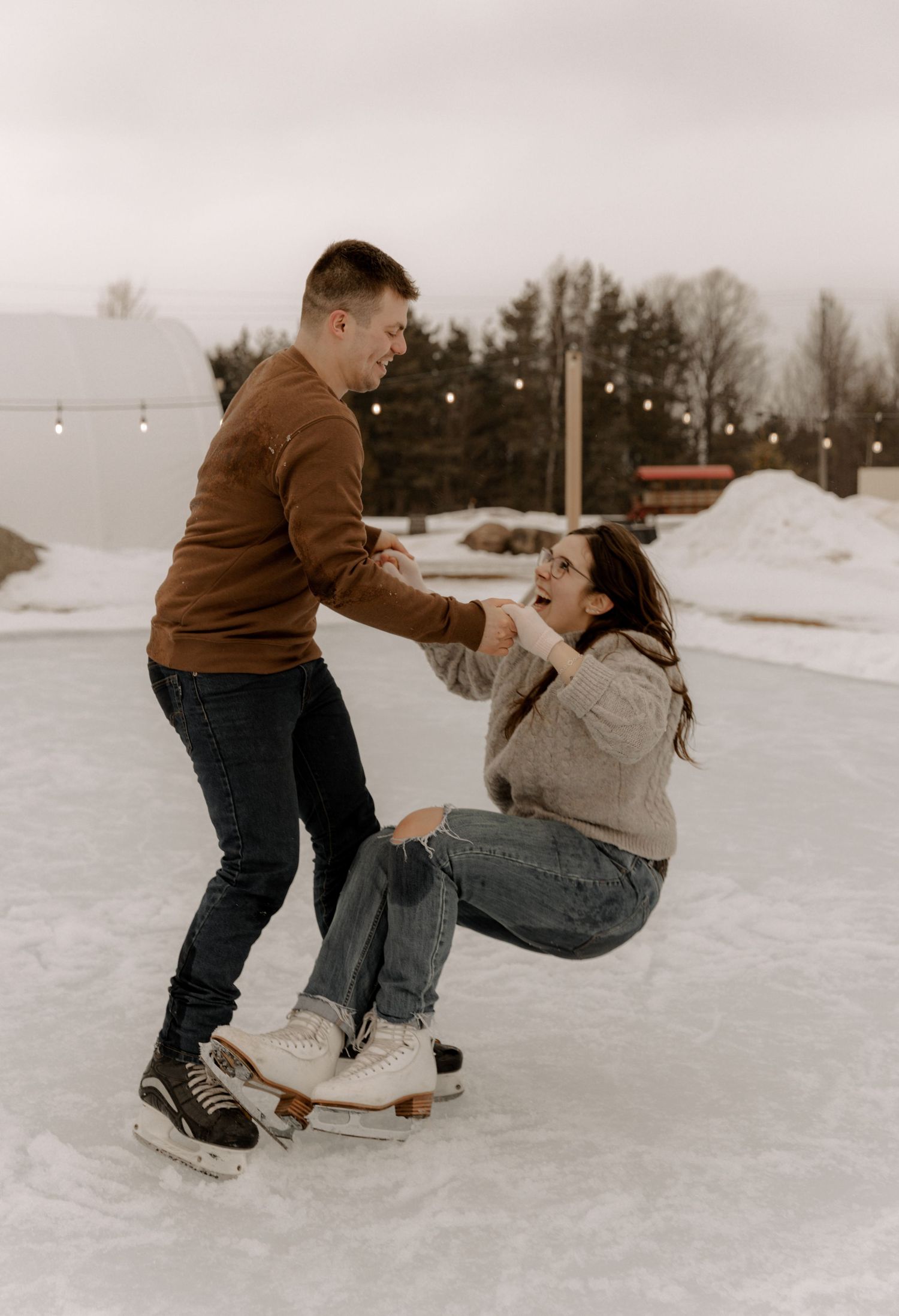 A couple ice skating together in winter, with string lights and snowy landscape in the background.