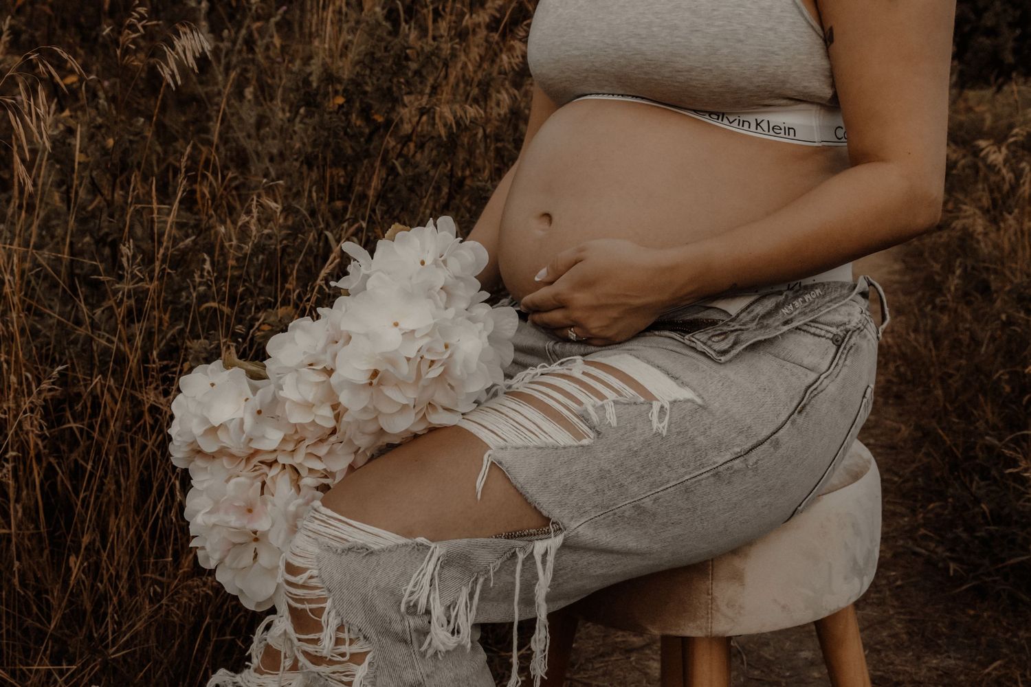 A pregnant person in distressed jeans holds a bouquet of white flowers while resting in a rustic outdoor setting.