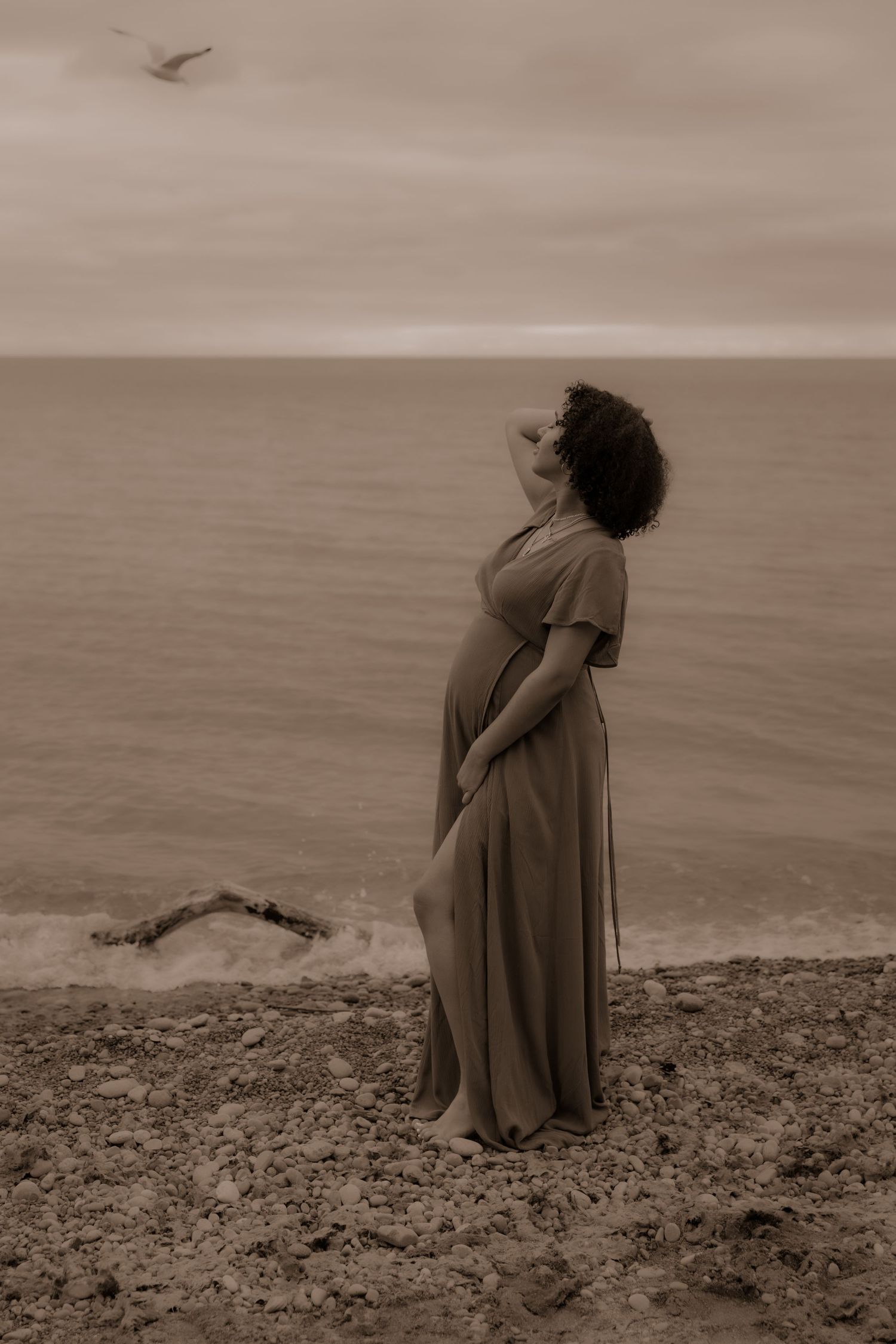 A sepia-toned photograph of an expectant mother in a flowing dress standing on a coastal cliff at sunset.