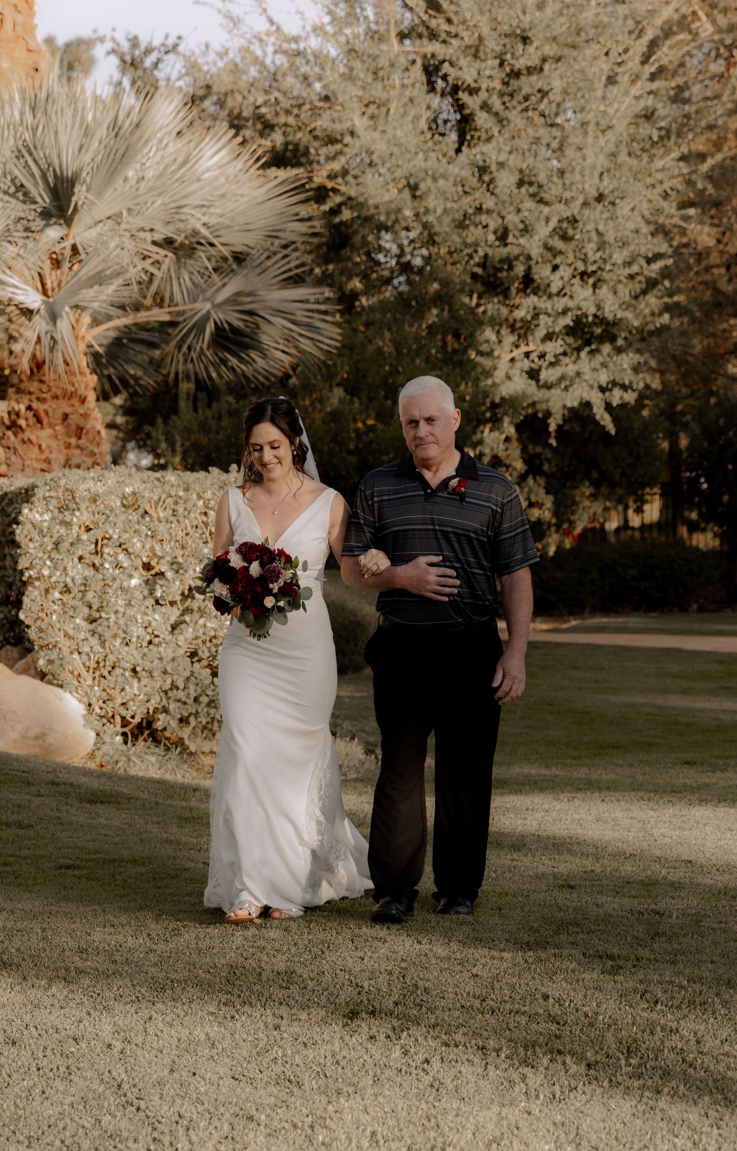 A wedding processional moment captured in warm sunlight with palm trees and garden landscaping.