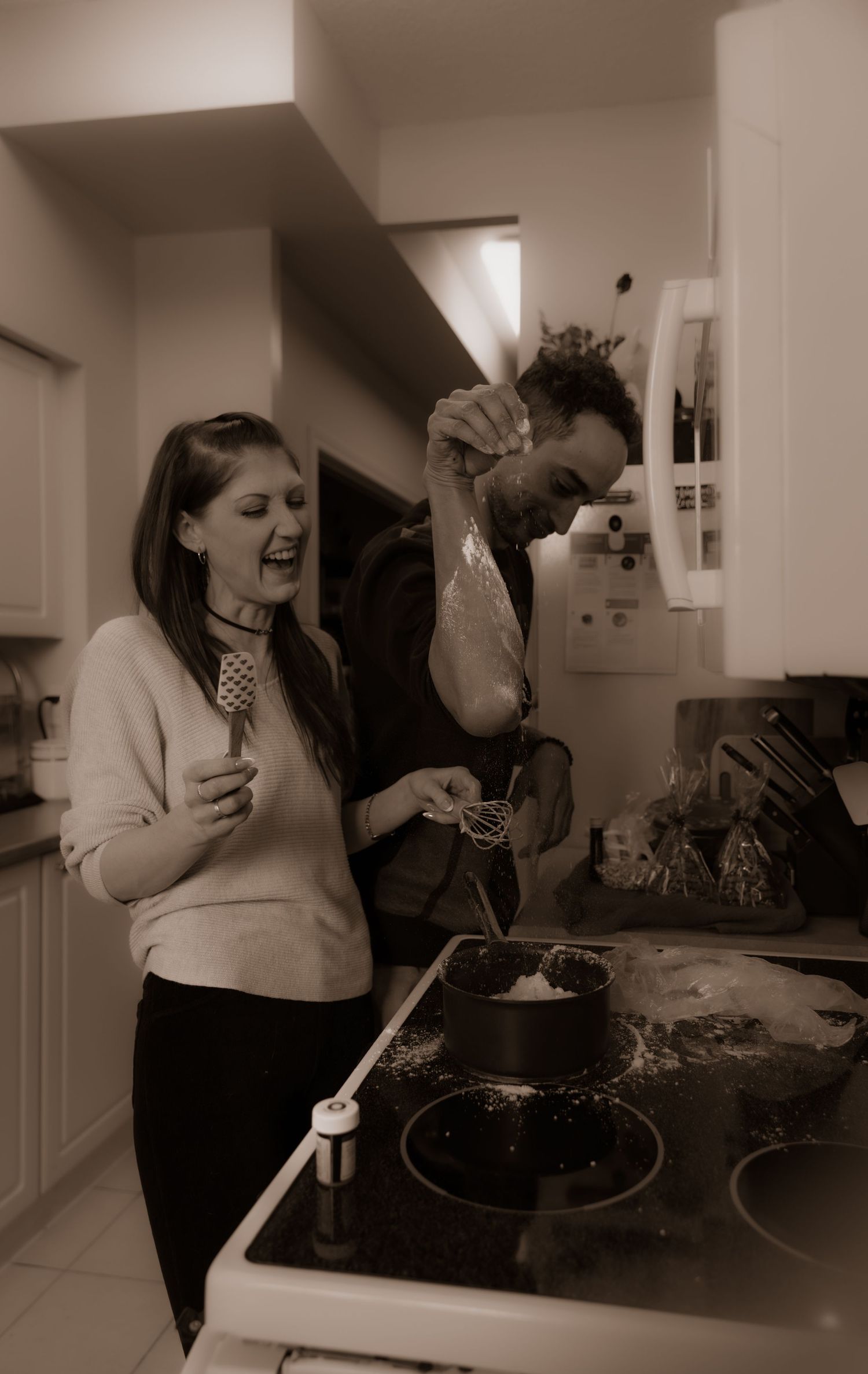 Two people laughing while cooking together in a kitchen with one person playfully holding ingredients.