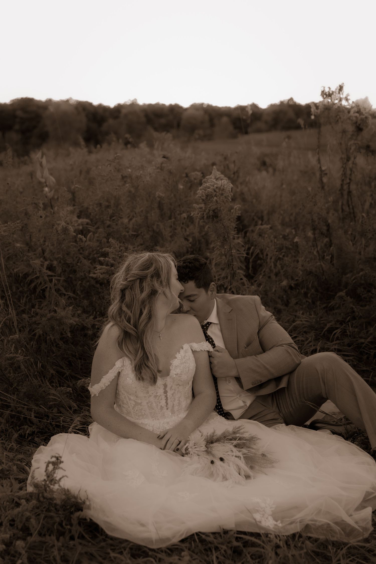 A romantic sepia-toned wedding photo of a couple sitting in a field at sunset.