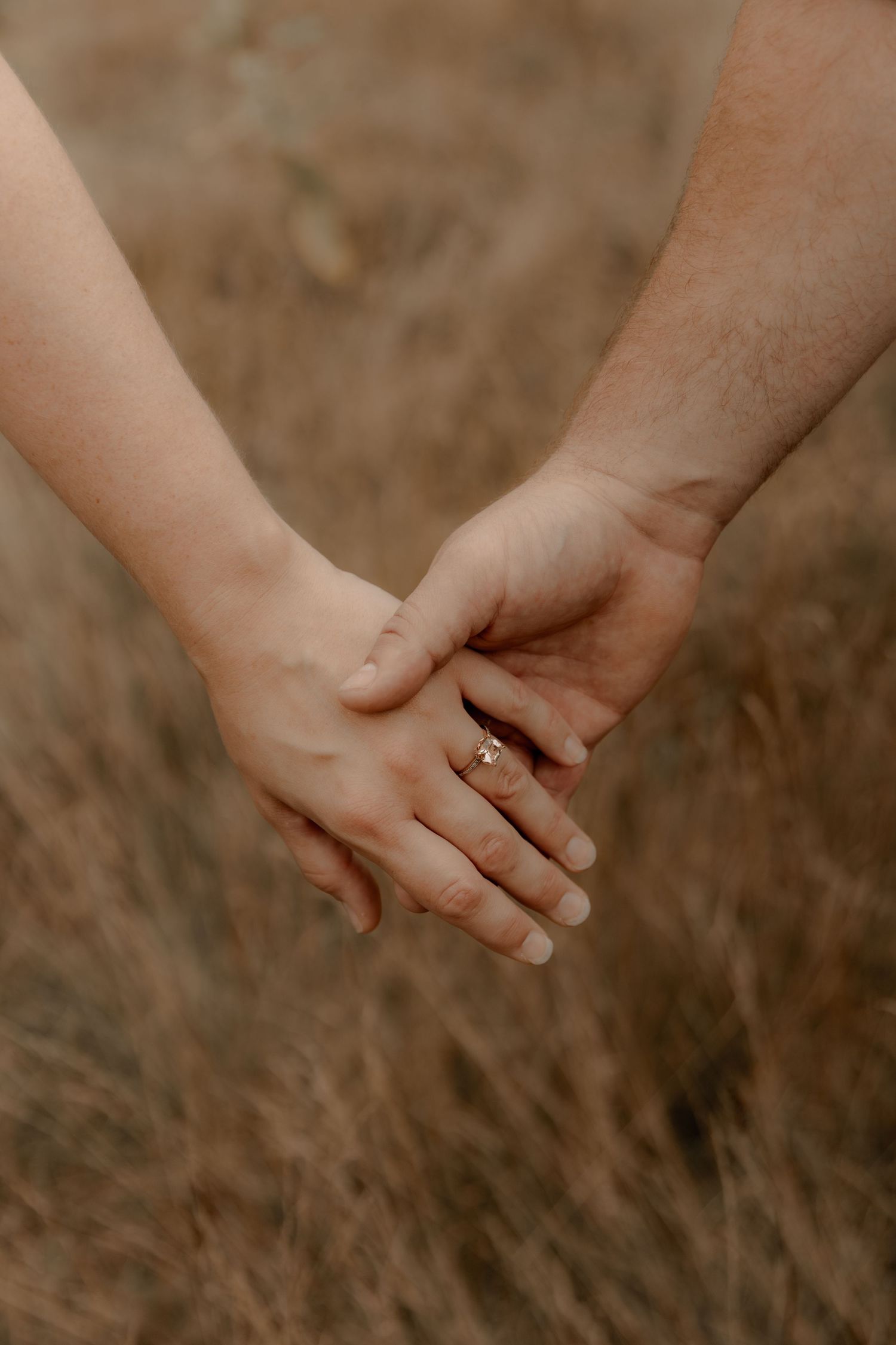Close-up shot of intertwined hands with engagement ring against a natural backdrop.