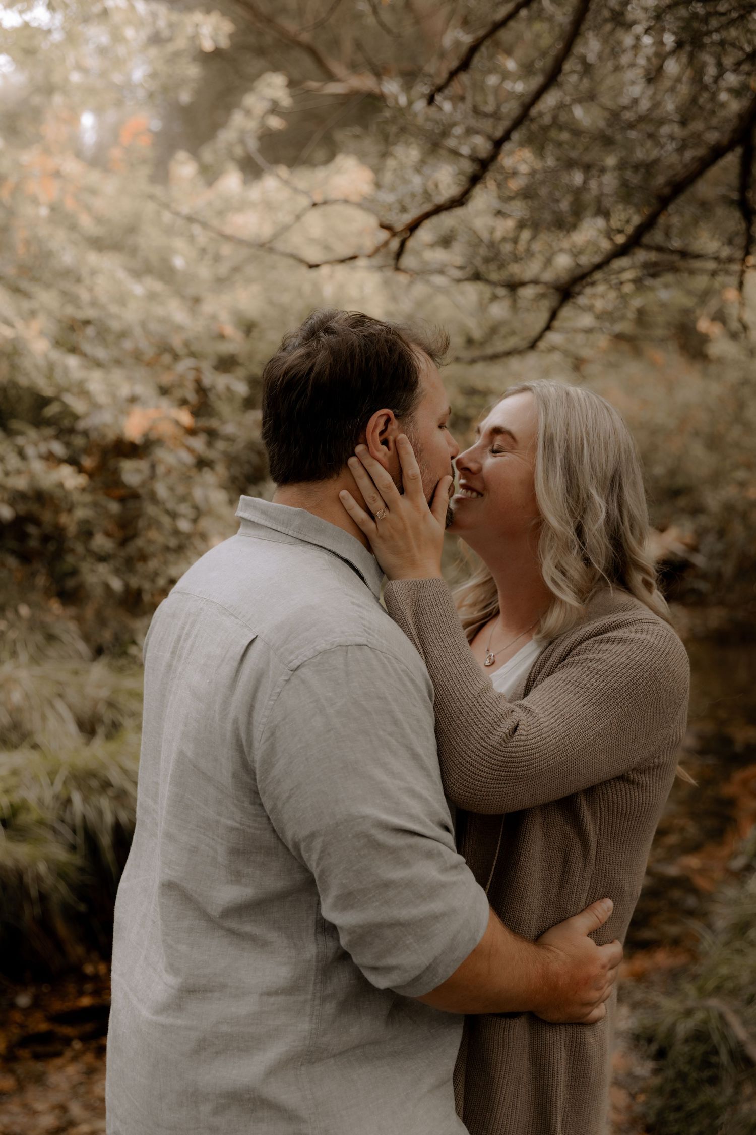 A romantic autumn engagement photo of a couple embracing in a dreamy forest setting with warm earth-toned colors.