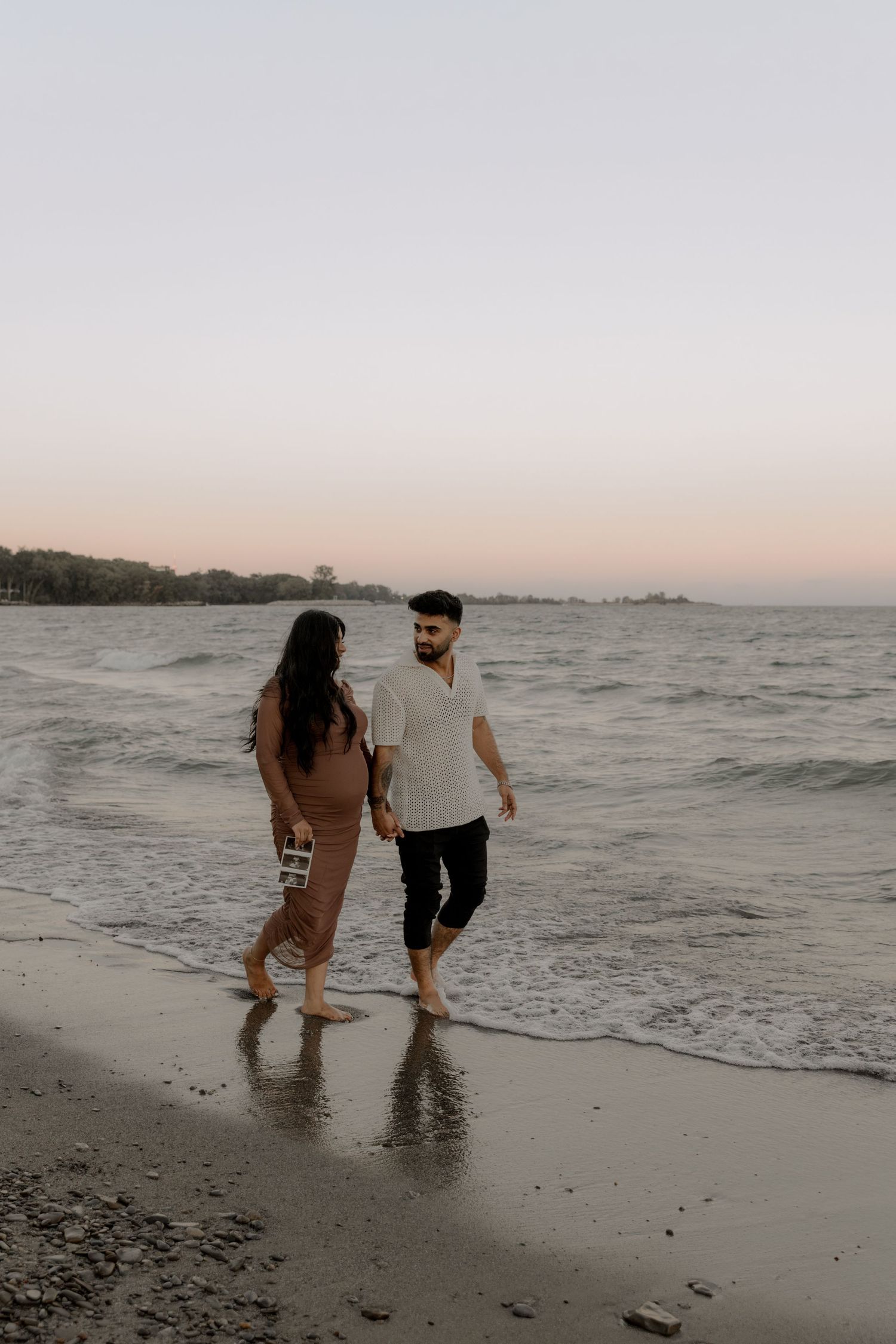 A couple walks together along the beach at sunset as waves gently lap at their feet.